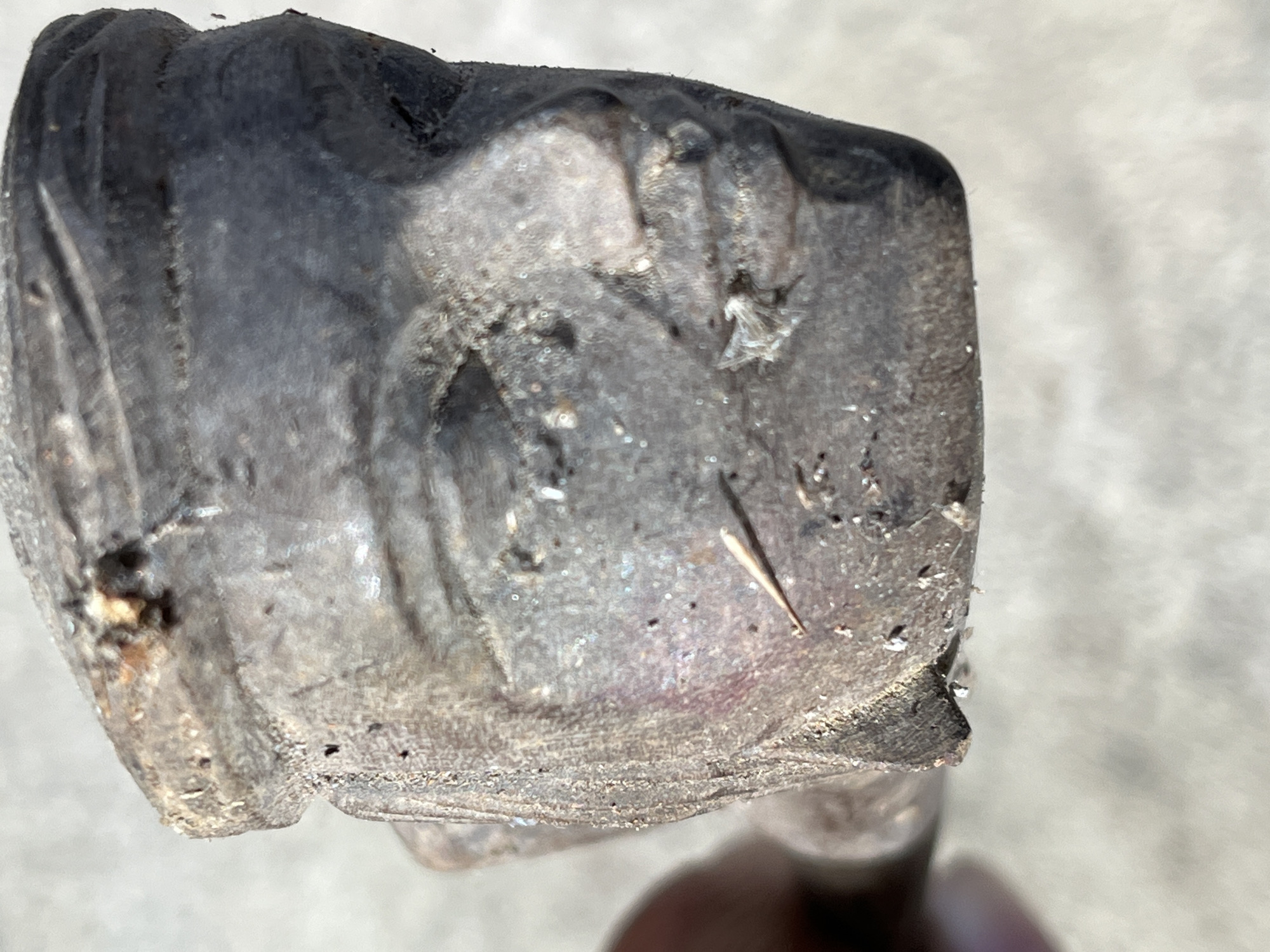 The image shows a close-up, forensic view of a weathered, dark-colored carved object, likely wood or bone, depicting a human face. The carving is rough and shows signs of age and wear, with visible chips, cracks, and accumulated debris. The face appears to have stylized features, including a visible forehead, eye sockets, nose, and lips. The object is held by a hand, partially visible in the lower left corner of the frame, suggesting it might be an artifact being examined. The background is out of focus and appears to be a light-colored, possibly paved surface, like concrete. No specific location in Tambakreja, Indonesia can be inferred from the image itself.