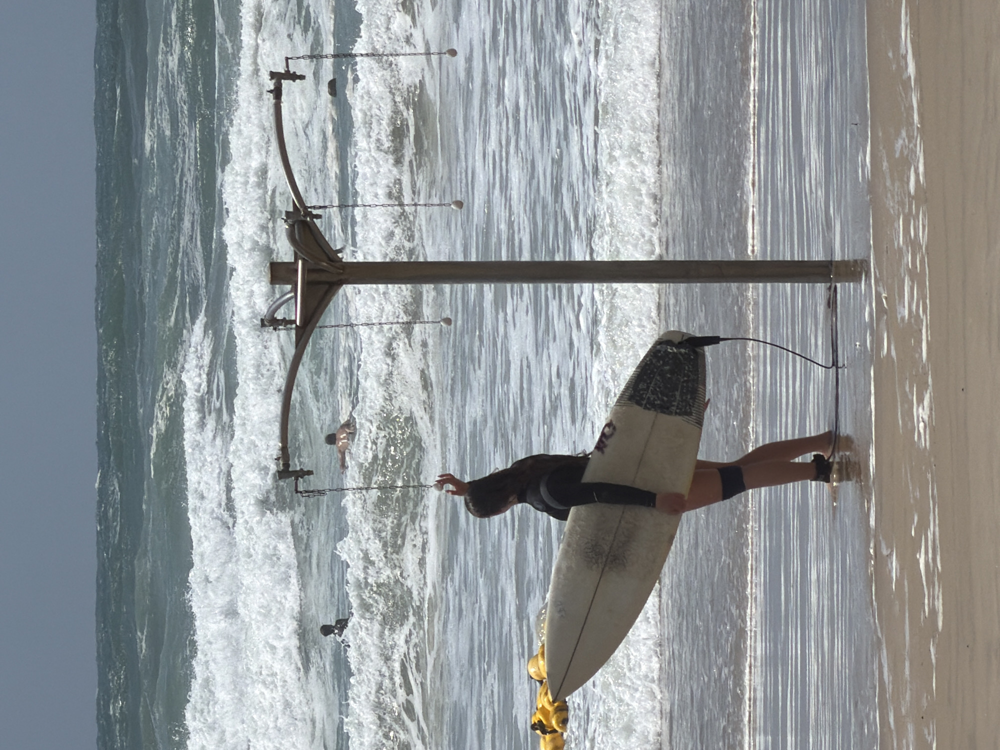 Here is a description of the image:
The image shows a beach scene. A person is holding a surfboard near a metal structure with chains and buoys. The person is on the sandy part of the beach, with the ocean waves visible in the background. Other people can be seen in the water. While the scene is consistent with a beach, there is not enough information in the image to confirm the exact location as Tel Aviv.