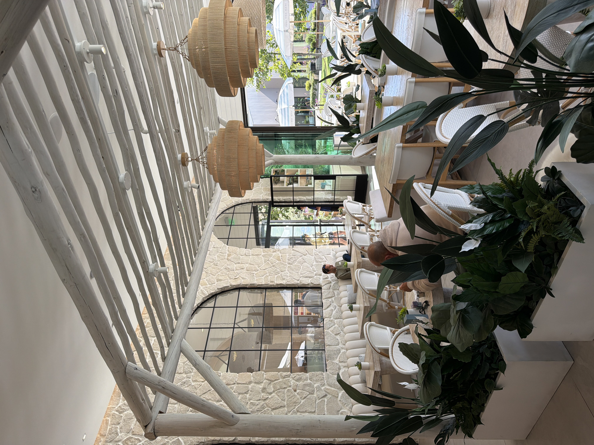 The image shows the interior of a restaurant with a relaxed, coastal vibe, likely located in Malibu, United States of America. The restaurant features a high ceiling with exposed wooden beams painted white, creating a rustic yet airy atmosphere. Two large, tiered pendant lights made of woven material hang from the ceiling, illuminating the dining area.
The walls are constructed from light-colored stone, adding to the natural and organic feel of the space. Large arched windows with black metal frames offer views of an outdoor seating area with white umbrellas and lush greenery. Inside, there are several dining tables with white wicker chairs. Some tables are set with white napkins and small potted plants as centerpieces.
In the foreground, large potted plants with dark green leaves are strategically placed, adding a tropical touch and defining seating areas. One man is seated at a table in the middle ground, facing away from the camera and looking down at his table. Another person's head is visible at the bottom right of the frame, also seated at a table. In the background, through the windows, several people are visible in the outdoor seating area. The overall impression is one of a bright, spacious, and upscale casual dining establishment.