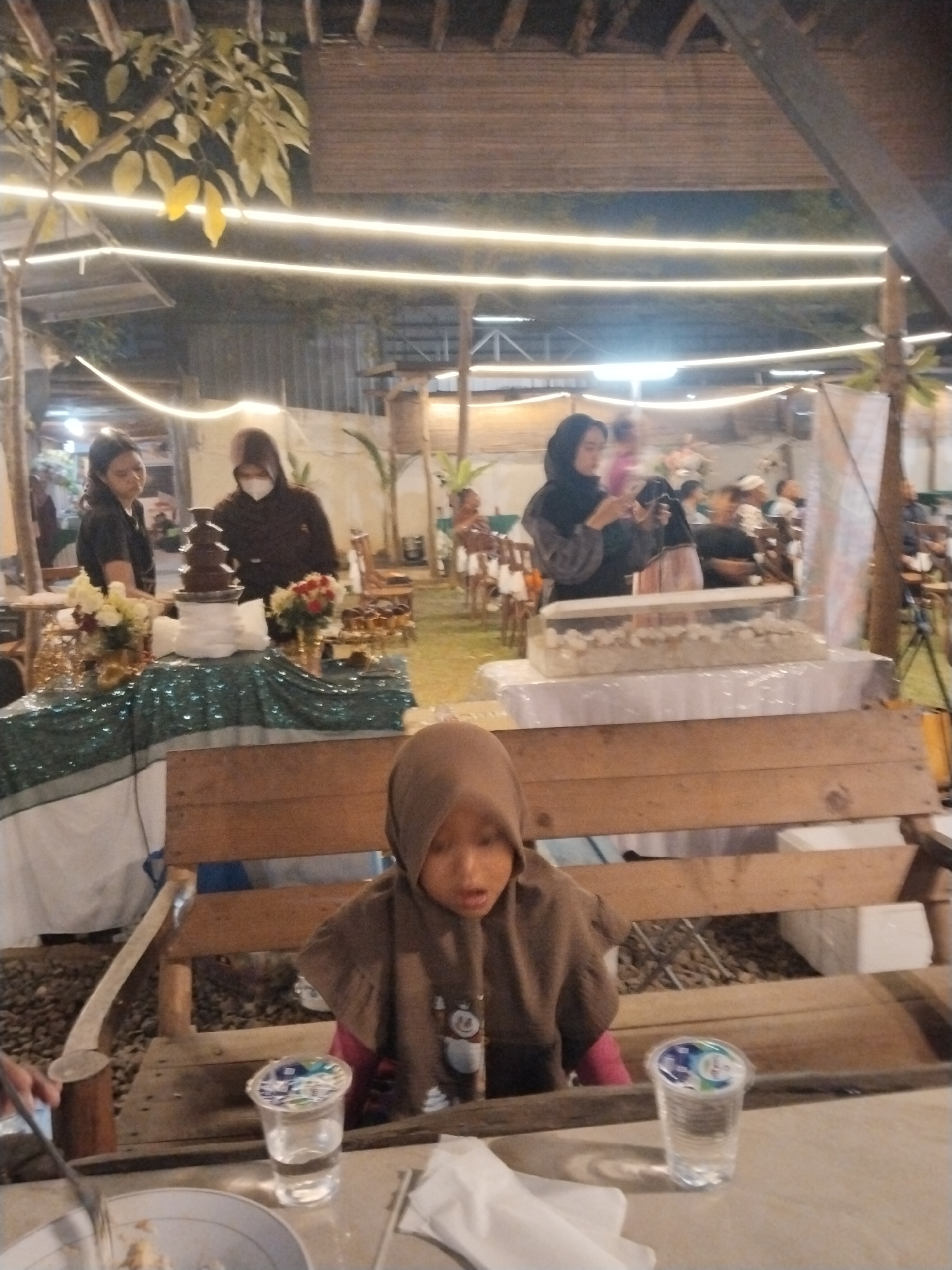 In Serang, Indonesia, an outdoor event is underway at night. The scene is illuminated by strings of white lights. In the foreground, a young child wearing a brown hijab sits at a wooden table, with a partially filled cup of water in front of them. Behind the child, another wooden bench is visible. Further back, a dessert table is set up with a chocolate fountain and floral arrangements. Several women are attending to the table, with one holding a phone. In the background, other people are seated at tables, and the event appears to be taking place in a garden or open-air venue.