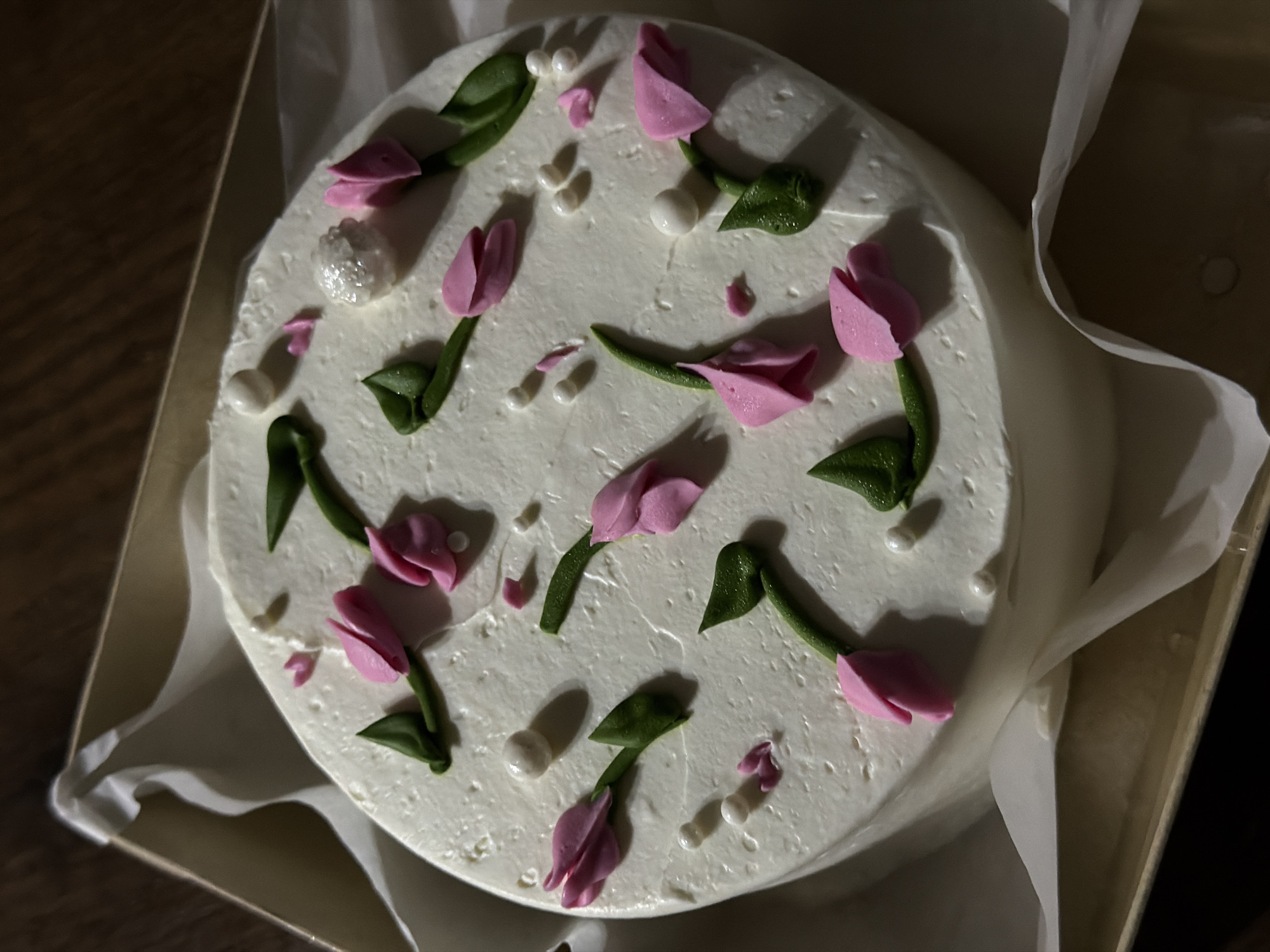 A top-down view of a small, round cake decorated with white frosting and pink tulip-like flowers with green stems and leaves. Small white pearl-like candies are scattered across the top. The cake is in a cardboard box lined with white paper. The setting appears to be indoors, with some wood visible around the edge of the box. There is no evidence to indicate this is located in Rossiyskiy, Russia.
