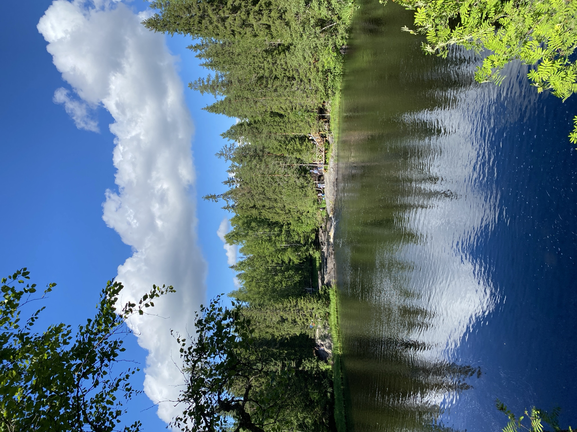 The image captures a serene natural landscape in Ruskeala, Russia. A dark, reflective body of water dominates the foreground, its surface rippled by gentle currents and reflecting the sky and surrounding trees. The water is bordered by a rocky shoreline and lush green grass.
In the mid-ground, a dense forest of coniferous and deciduous trees forms a vibrant backdrop. The trees are tall and densely packed, with their green foliage creating a rich texture.
A wooden boardwalk and bridge are visible along the shoreline, with several people casually walking on them or standing near the water's edge. These individuals appear to be tourists or visitors enjoying the scenery. Some are gathered in small groups, while others are more spread out.
The sky above is a brilliant blue, dotted with fluffy white clouds that are casting soft shadows on the landscape. The overall impression is one of a peaceful, picturesque natural park. The location is likely part of the Ruskeala Mountain Park, known for its marble canyons and surrounding forests.