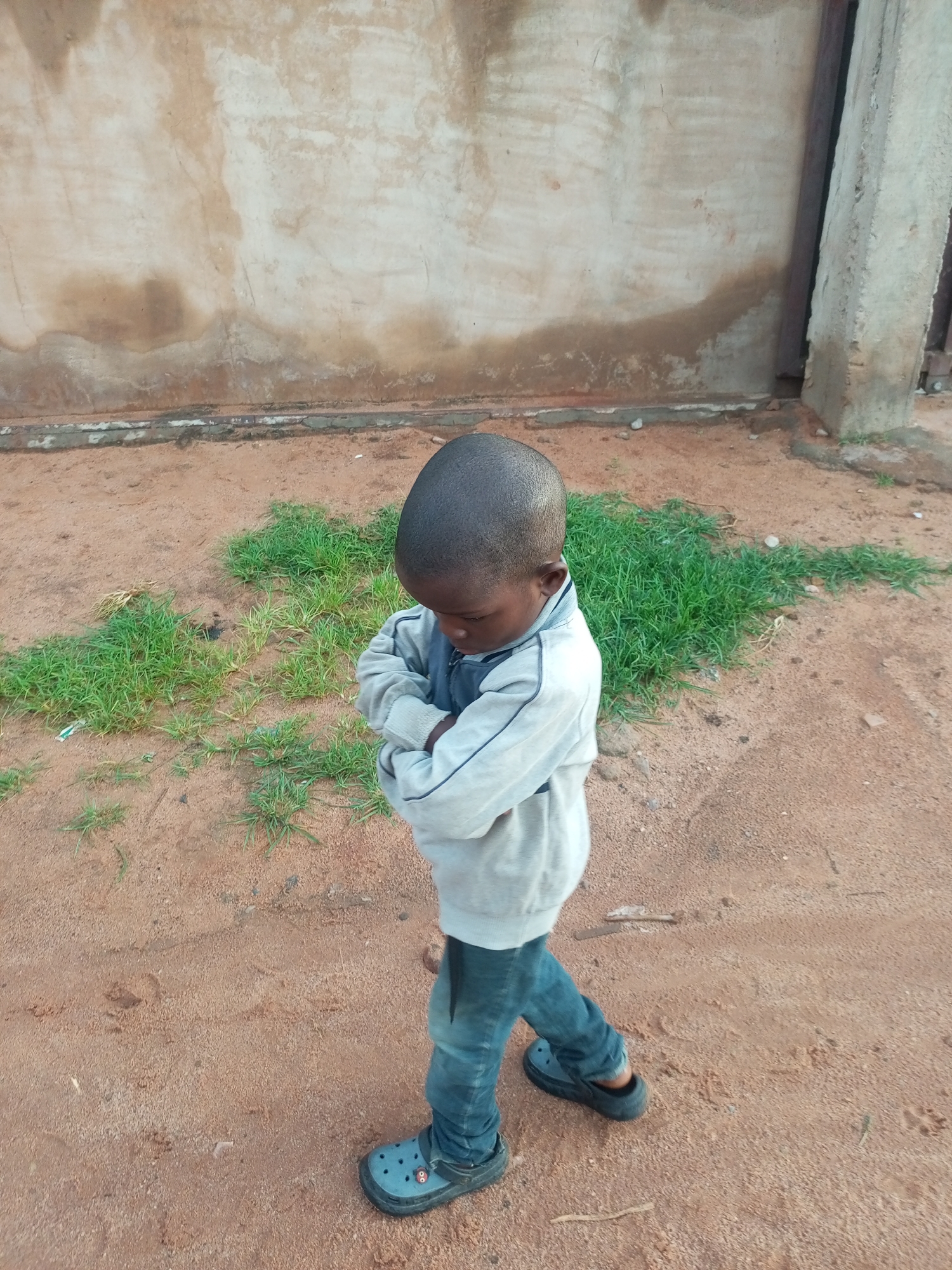 A young Black boy is shown from the chest down, walking to the right with his arms crossed and head down, in an outdoor setting. He is wearing a light gray sweatshirt with dark gray accents and blue jeans, and dark blue Crocs-style shoes. The ground is sandy and reddish-brown, with patches of green grass scattered throughout. In the background, a textured, light brown wall runs horizontally, with a concrete pillar on the right side. The scene appears to be in an undeveloped or rural area within Tempure, Nigeria.