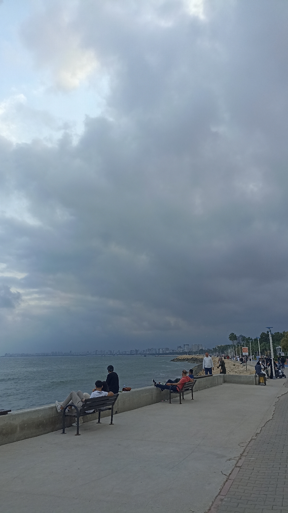 The image depicts a waterfront scene in Yenişehir, Turkey, on a cloudy day. People are relaxing on benches along a concrete walkway that borders the sea. In the distance, a skyline of buildings is visible across the water. The sky is overcast with dark clouds. Some people are walking along the promenade, while others are seated, facing the sea. A low concrete wall separates the walkway from the water's edge. Palm trees and lampposts line the walkway.