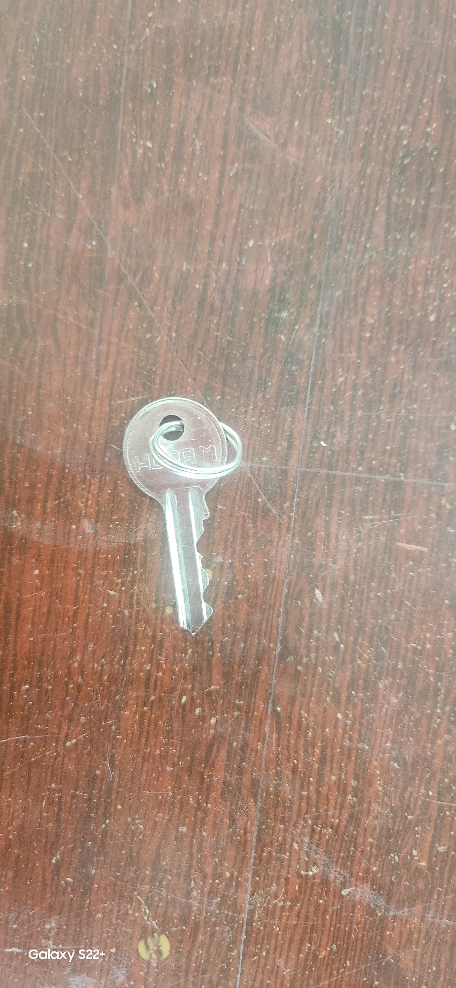 A silver metal key with a circular bow is placed on a wooden surface. The bow of the key has faint markings that appear to be letters, possibly "HAN M". The key is oriented diagonally across the frame, with the bow at the top left and the teeth pointing towards the bottom right. The wooden surface is dark brown with a visible wood grain pattern and appears somewhat worn with small flecks of debris scattered across it. A faint, almost imperceptible line runs vertically down the center of the frame. The bottom left corner of the image displays the text "Galaxy S22+". There is no discernible context regarding the city of Nyurba, Russia, within the visual information provided.