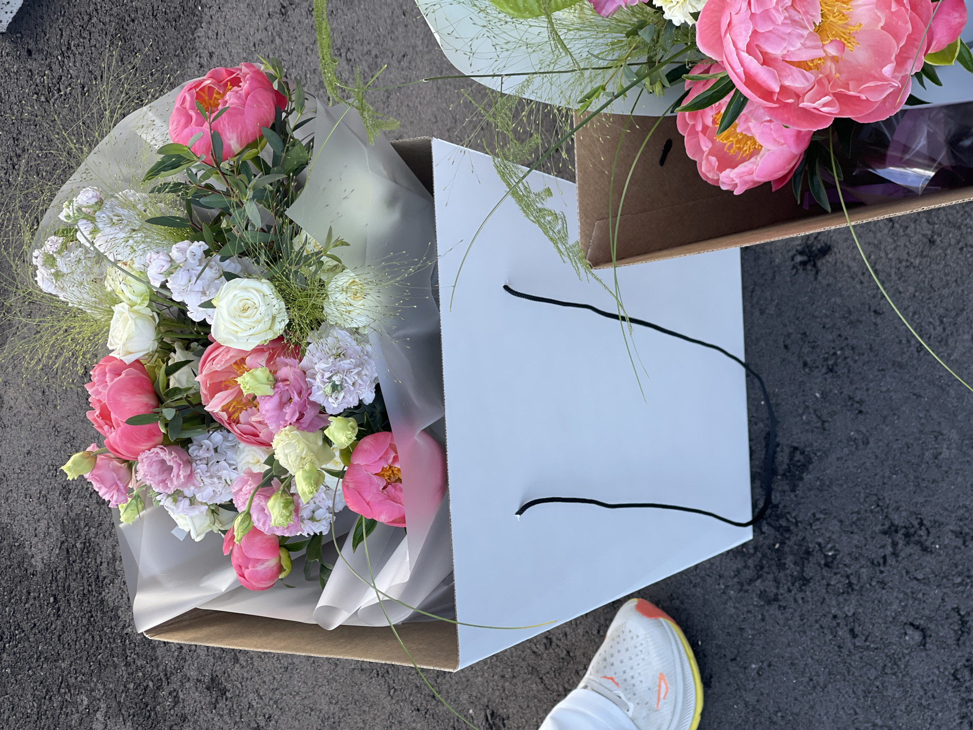 Two gift bags containing floral arrangements are positioned on an asphalt surface. The bag on the left is white with black handles and holds a bouquet of pink and white peonies, roses, and other mixed flowers. A white sneaker with yellow and orange accents is visible to the left of this bag. The bag on the right is made of brown cardboard and contains a bouquet of pink peonies and other green foliage. The scene appears to be outdoors.