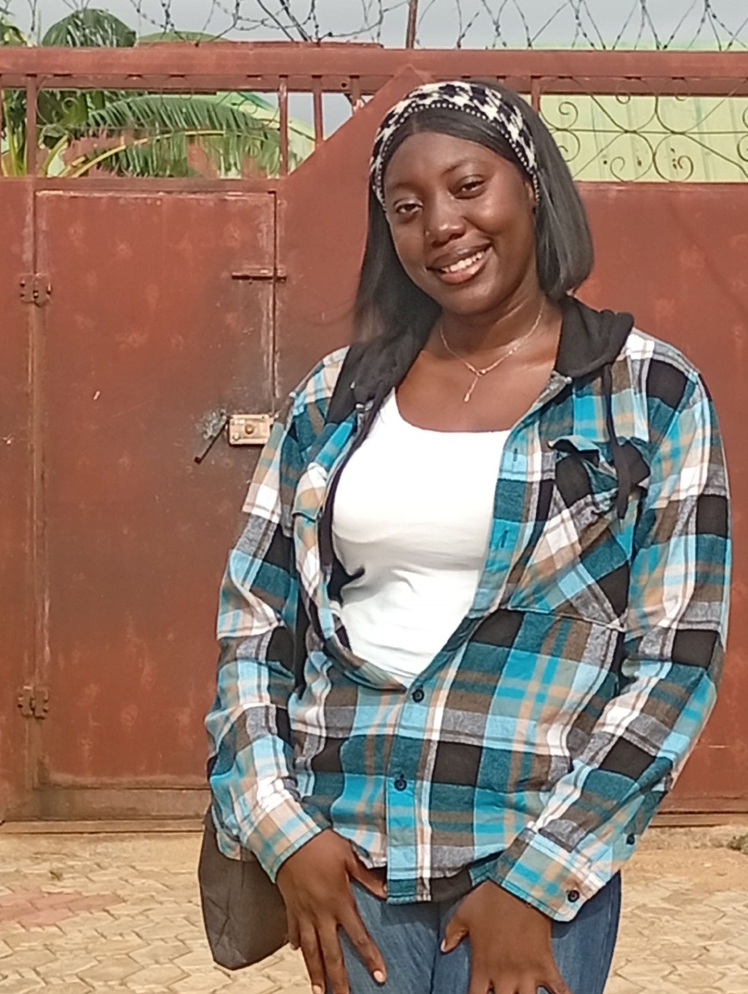 A young woman stands outdoors in Rido, Nigeria. She is smiling and wearing a white t-shirt, an open blue and brown plaid shirt with a black hoodie underneath, blue jeans, and a black and white patterned headband. She also has a necklace on. Behind her is a large, red metal gate topped with barbed wire. A plant with broad green leaves can be seen through the gate. The ground is paved with reddish-brown bricks.