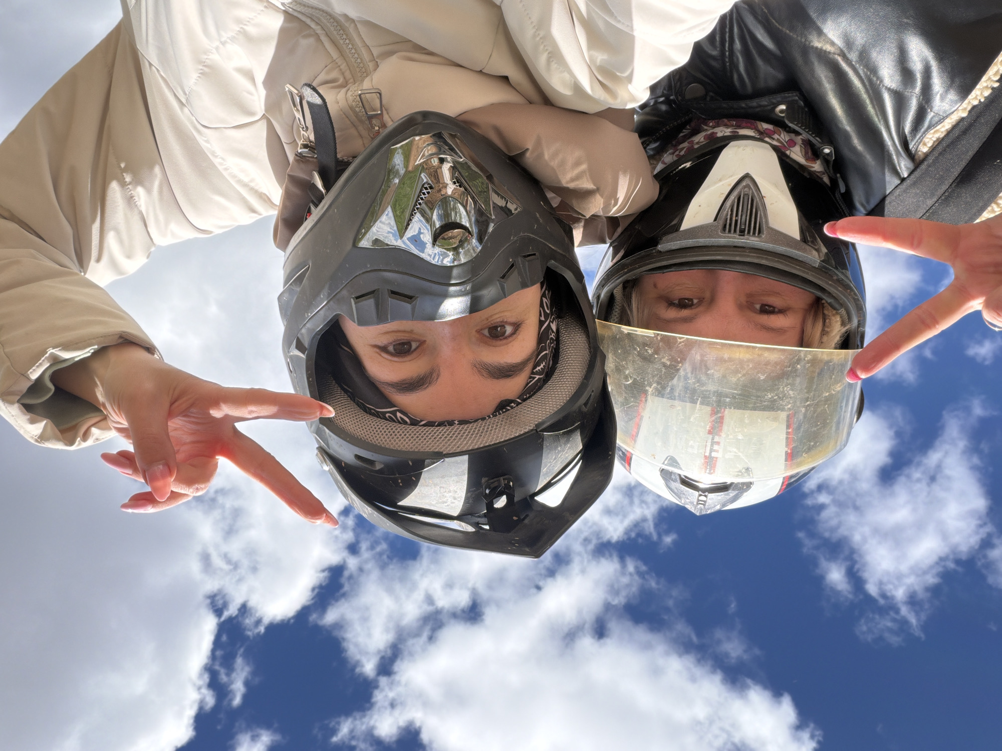 Two people wearing motorcycle helmets are angled upside down toward the viewer. The person on the left is wearing a tan puffy jacket, and the person on the right is wearing a black leather jacket. Both individuals are making peace signs with their hands. The background is a blue sky with scattered white clouds. It is not possible to determine the location as Nizhnii Vishur, Russia from the given information.