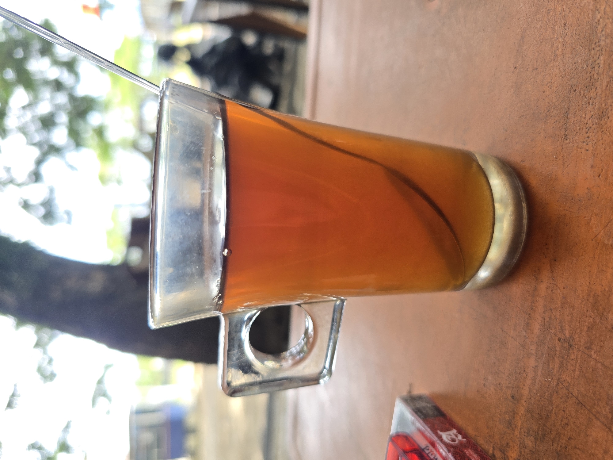 In the city of Meunasah Pupu, Indonesia, a clear glass mug filled with amber-colored tea sits on a wooden table. A silver spoon is partially submerged in the tea. To the left of the mug, a pack of cigarettes rests on the table. The background is blurred, showing a tree trunk, foliage, and a hint of a vehicle. The overall scene suggests an outdoor setting, possibly a cafe or roadside eatery.