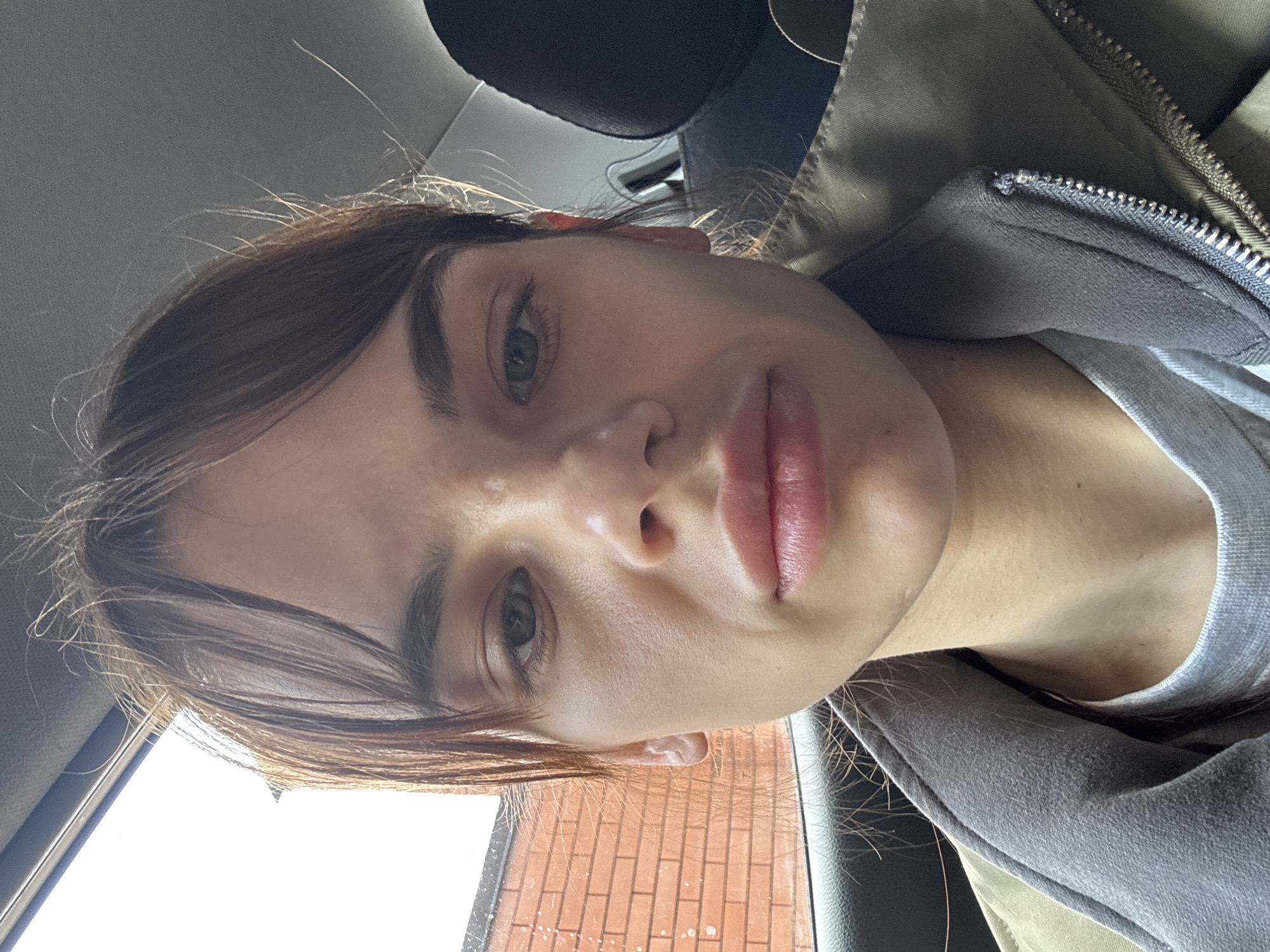 A close-up selfie of a fair-skinned woman with brown hair and green eyes. She is wearing a gray sweatshirt under a zip-up jacket. The background suggests she is in a car; a headrest is visible and a brick wall is seen through the window. There is no evidence to suggest that the location is Rostov-on-Don, Russia.