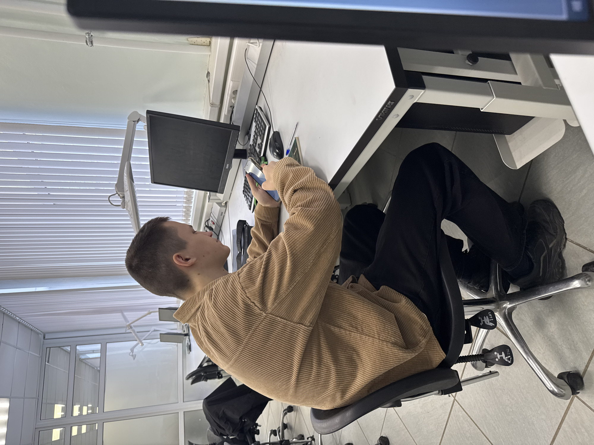 This media depicts a young male in an indoor environment, likely an office, lab, or classroom, located in Perm, Russia. The subject is seated at a white desk in a black office chair with wheels. He is wearing a light brown corduroy long-sleeved shirt, dark pants, and dark shoes, and has short, dark hair. He is holding a smartphone with both hands, appearing focused on its screen.
The desk setup includes a computer monitor, a keyboard, a computer mouse, a blue pen, and a notebook. A white power strip with electrical outlets is mounted on the wall behind the desk. The brand "VIKING" is visible on the side of the desk frame. The office chair features levers for adjustment, with the word "LIFT" and other icons clearly visible on them.
The room features white walls and a white tiled floor. A large window with horizontal white blinds is seen to the left of the subject, admitting natural light. Further in the background, there is a glass partition or door. A flexible white lamp arm extends from the upper part of the wall or ceiling, and a small, ceiling-mounted fixture, possibly a fire sprinkler head, is also present. The overall lighting suggests daytime. The subject's expression is neutral, indicative of concentration.