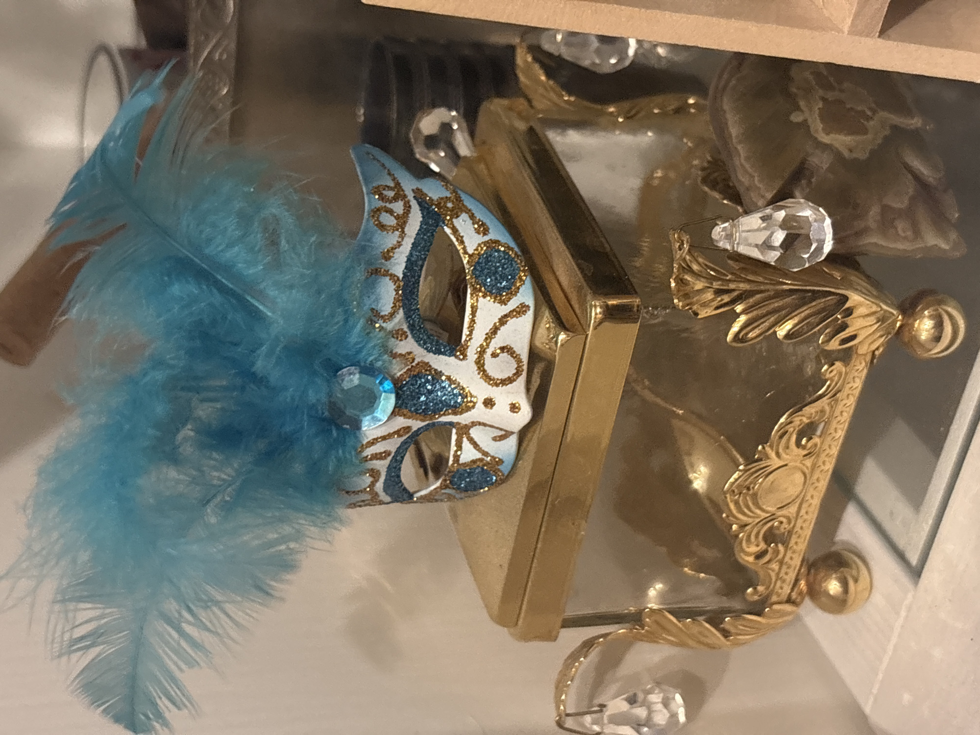 The image shows a decorative masquerade mask with blue feathers and glitter, resting on a golden, ornate glass box. The box has decorative gold trim and crystal pendants hanging from its sides. The scene appears to be an indoor setting, likely a display or shelf. The backdrop includes out-of-focus elements that suggest other decorative items or shelves. No people or actions are depicted in the image.
