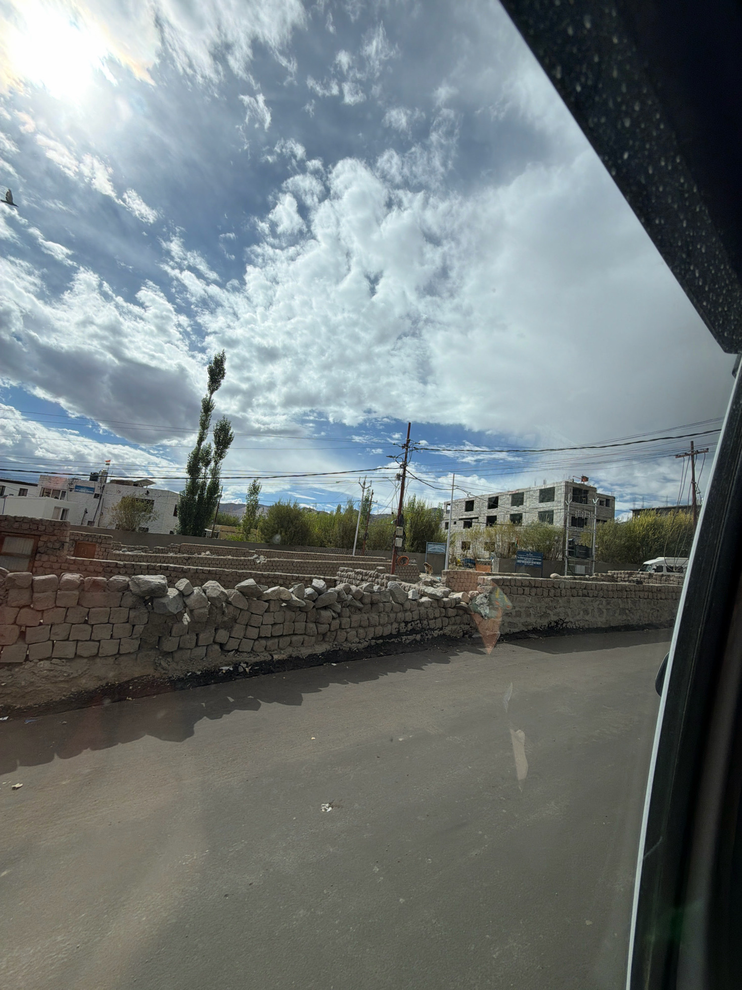 The image depicts a view from inside a vehicle looking out towards a street scene in Leh, India. The sky is partly cloudy with bright sunshine filtering through. A stone wall runs along the side of the road, with a paved surface in the foreground. In the background, there are buildings, trees, and utility poles. One building appears to be under construction. There are no people clearly visible in the scene. The overall impression is of a somewhat arid, mountainous region.