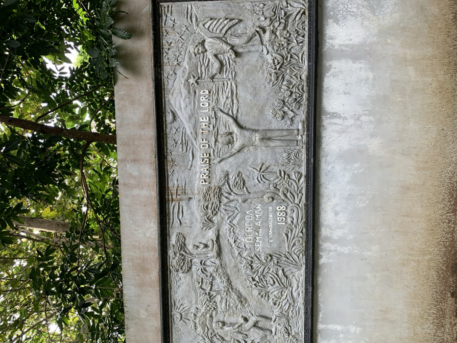A gray concrete wall with a bas-relief sculpture depicting a scene of praise and worship. The sculpture is set within a black frame and features a man with his arms raised to the heavens. In the background, there is a mountain with a crescent moon in the sky, and another figure with wings holding a staff with a star on top. Below the sculpture are the words "Praise of the Lord," "Berdoa Semalaman," and "1 Tawarikh 28:10 1988" in white. The wall is situated in a forested area, with tree branches and leaves visible above it. The location is in Kecamatan Borobudur, Indonesia, based on the coordinates -7.606 latitude and 110.181 longitude.