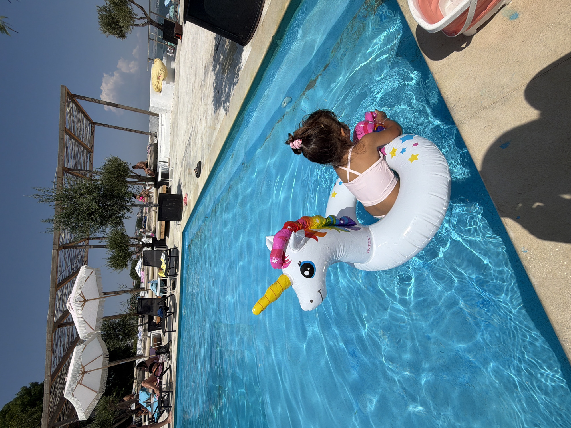 A young girl, with her hair tied up in a pink scrunchie, sits in a bright blue swimming pool. She is wearing a pink tank top. She is on a white inflatable unicorn floatie with a rainbow mane and a yellow horn. She is holding a pink toy in her hands. The pool is surrounded by a light-colored deck. In the background, there is a wooden structure with a thatched roof, green trees, lounge chairs, and umbrellas. The sky is blue with a few white clouds. The scene appears to be set on a sunny day, likely during the daytime. The overall mood is one of leisure and enjoyment.
There is visible text on the inflatable floatie that reads "INTEX". The location context provided is Burgas, Bulgaria.