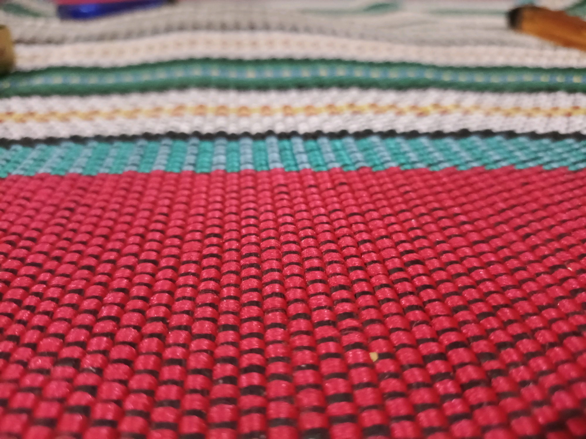 This is a close-up, shallow depth-of-field photograph of a woven mat. The mat features horizontal stripes of various colors including red, teal, white, green, and a light beige with yellow accents. The texture of the weave is clearly visible, with raised red threads forming rows interspersed with darker, recessed threads. The closest stripe is a vibrant red, which dominates the foreground and is in sharp focus. The rest of the mat recedes into a blur of color. The image does not depict any specific location within Rancang Kencono, Indonesia, or any people, objects, actions, or interactions.