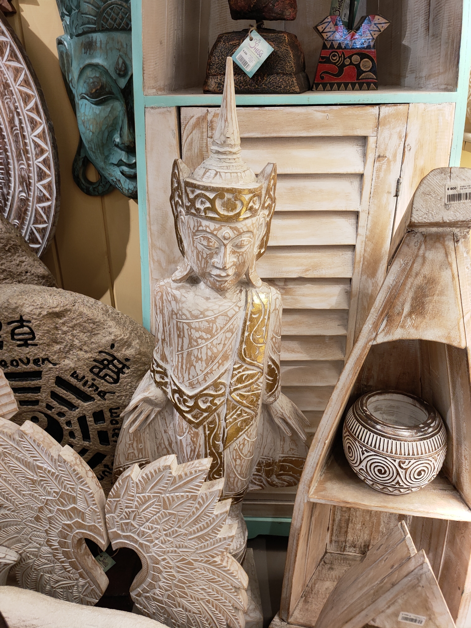 The image displays a still life arrangement of various decorative items, primarily carved wood, suggesting an indoor setting such as a shop or home decor display. The central subject is a standing Buddha statue, intricately carved with a serene expression. It is adorned with gold detailing on its robe and crown, and its body exhibits a distressed white finish.
To the left of the Buddha, a large, rough-hewn stone is visible, inscribed with black calligraphy-like symbols. Further to the left and slightly behind, a turquoise-colored carved mask with a large headdress is partially visible, adding an element of ethnic or cultural decor. In the foreground, two carved wooden wing-like objects are positioned to form a heart shape.
To the right of the Buddha, a rustic wooden cabinet with louvered doors forms the backdrop. A shelf above the louvered doors holds several smaller decorative pieces. One is a dark, textured object with a tag, possibly a lamp base, and another is a colorful, abstract wooden carving with geometric patterns. On a lower shelf of the cabinet, a round, textured bowl with a white spiral pattern is displayed within a triangular wooden structure.
The overall aesthetic is one of handcrafted, artisanal decor, with a focus on natural materials and intricate carving. The lighting appears to be indoor ambient lighting, with no indication of a specific time of day or weather conditions. There is no discernible emotional tone, as the scene focuses on the objects themselves.
Visible text includes price tags on some of the items. One tag on the dark object has "R230" and another on the wooden bowl has "6 900". A tag on the carved wings reads "Sweet". The setting is identified as Sochi, Russia, which provides geographical context but no visual cues within the image itself.