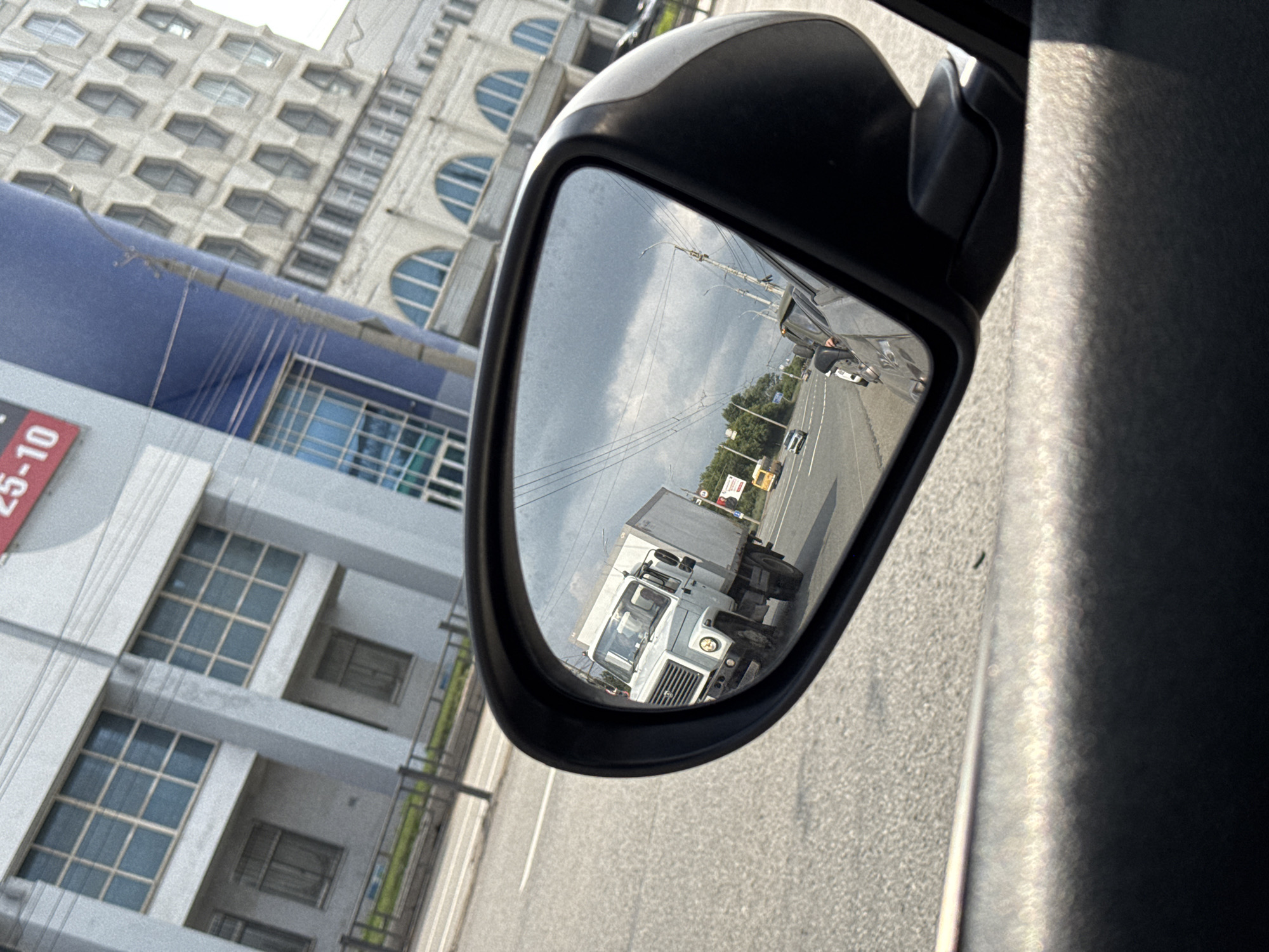 The image is a photograph taken from inside a vehicle, looking into a side-view mirror. The mirror reflects a road with several vehicles, including a white truck in the foreground. The road appears to be an urban street with trees and buildings lining one side. In the background, a multi-story building with a patterned facade and arched windows is visible. To the left of the mirror, parts of the vehicle's interior and the exterior of a building are seen. A red sign with white text that reads "25-10" is partially visible on the building. The scene suggests daytime with overcast skies, as indicated by the cloud cover in the reflection. The overall impression is a snapshot of a moment while driving through a city.