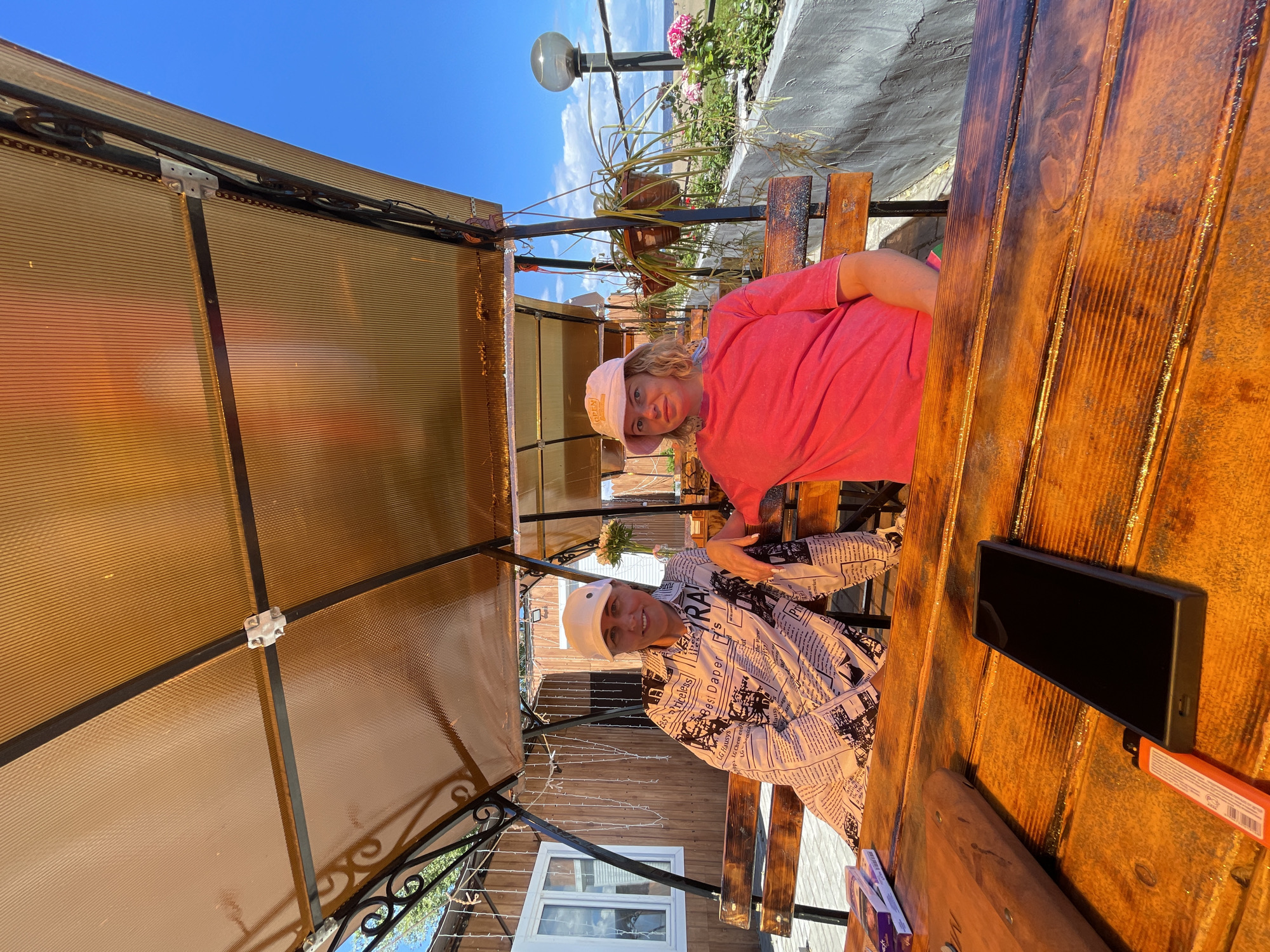 Two women are sitting on a wooden bench under a translucent amber-colored awning in an outdoor seating area. The woman on the left is wearing a white cap and a long-sleeved shirt with a newspaper print. The woman on the right is wearing a pink bucket hat and a bright pink t-shirt. They are seated at a wooden table. On the table are a mobile phone, a lighter, and a stack of cigarette packs. Behind them, there is a wooden building with string lights, potted plants, and a view of the sea and a beach in the distance. The sky is clear and blue with some clouds. This scene is located in Belozersk, Russia, likely at a cafe or restaurant with an outdoor seating area.