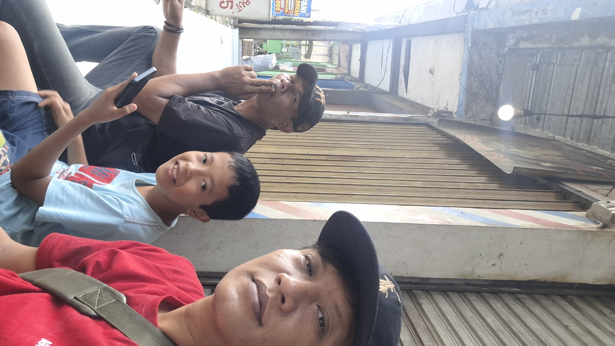 A selfie is taken from the perspective of a person in the foreground on the right, who is wearing a red shirt and a black baseball cap. The person is looking towards the camera with a neutral expression. To the left of the foreground person is a young boy wearing a light blue t-shirt and dark blue shorts, smiling at the camera. Further to the left is an adult male wearing a black t-shirt and a black baseball cap, with his index finger near his mouth, appearing to be smoking or contemplating. He is holding a black mobile phone. They are all seated next to a building with a corrugated metal roll-up door. The building has a concrete pillar with red and blue stripes, characteristic of a barber shop. A light bulb hangs from the ceiling above them. In the background, a sign with Indonesian text and some white markings is visible. The environment suggests an outdoor setting in an urban area, possibly Cilaku, Indonesia, given the context.
