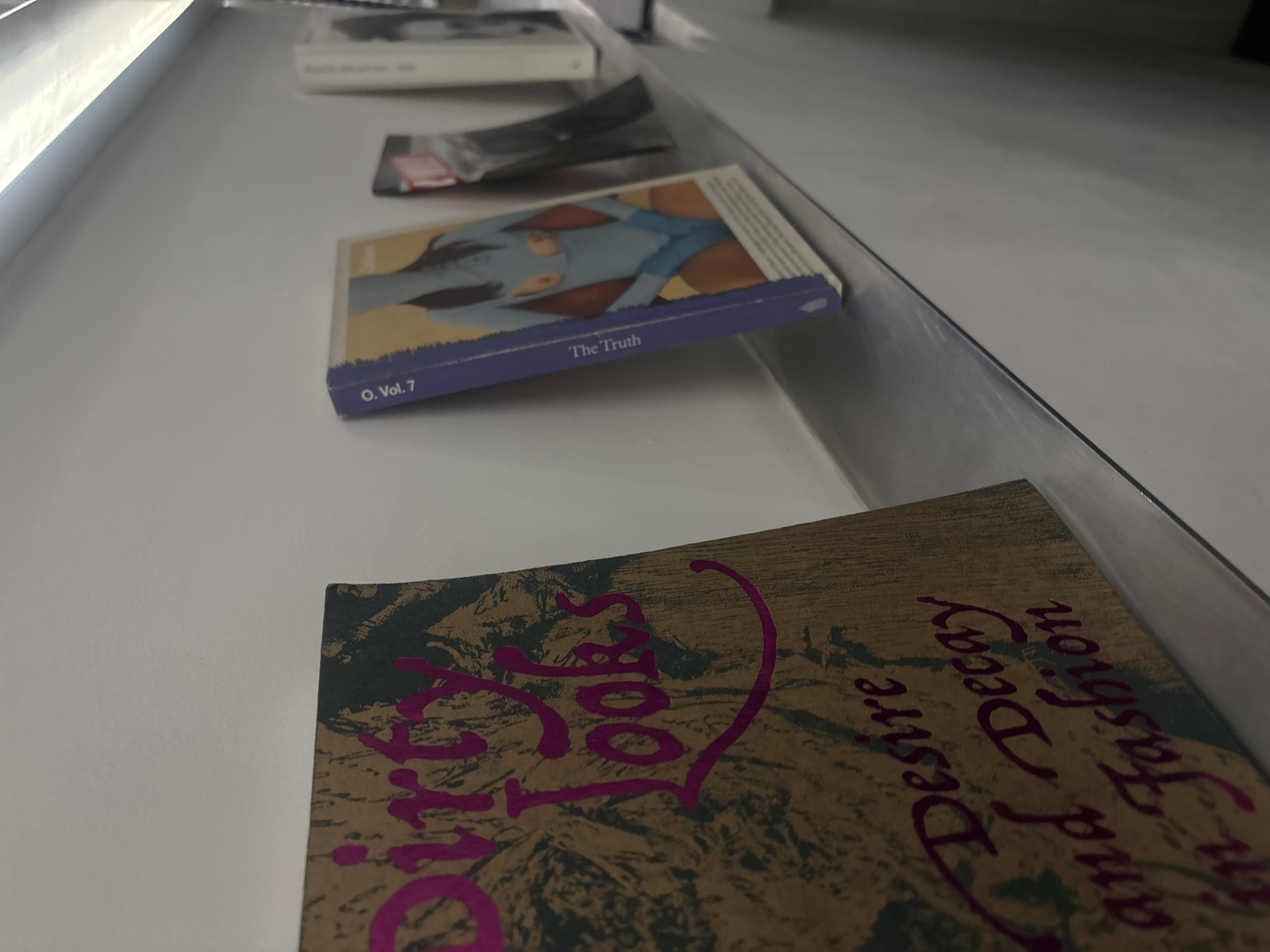 The photograph presents a close-up, high-angle view of a display of books in what appears to be a gallery or bookstore setting, possibly in Mexico City, Mexico, given the content of some of the book titles. The books are arranged on a horizontal, white surface, seemingly a shelf or display table, with a reflective edge suggesting the presence of glass or a similar material.
Starting from the foreground and moving into the depth of field, the most prominent book is titled "Dirty Looks: Desire, Decay and Fashion," featuring a cover with stylized imagery and distinct pink lettering. Behind it, another book titled "The Truth, O. Vol. 7" is visible, showcasing an illustration of a woman in blue. Further back, partially obscured, is a book with a silver and black cover. In the background, a slim white book is lying horizontally. The lighting is bright and appears to be artificial, highlighting the book covers. The lack of human presence suggests the image was taken in a moment when the display area was unoccupied.