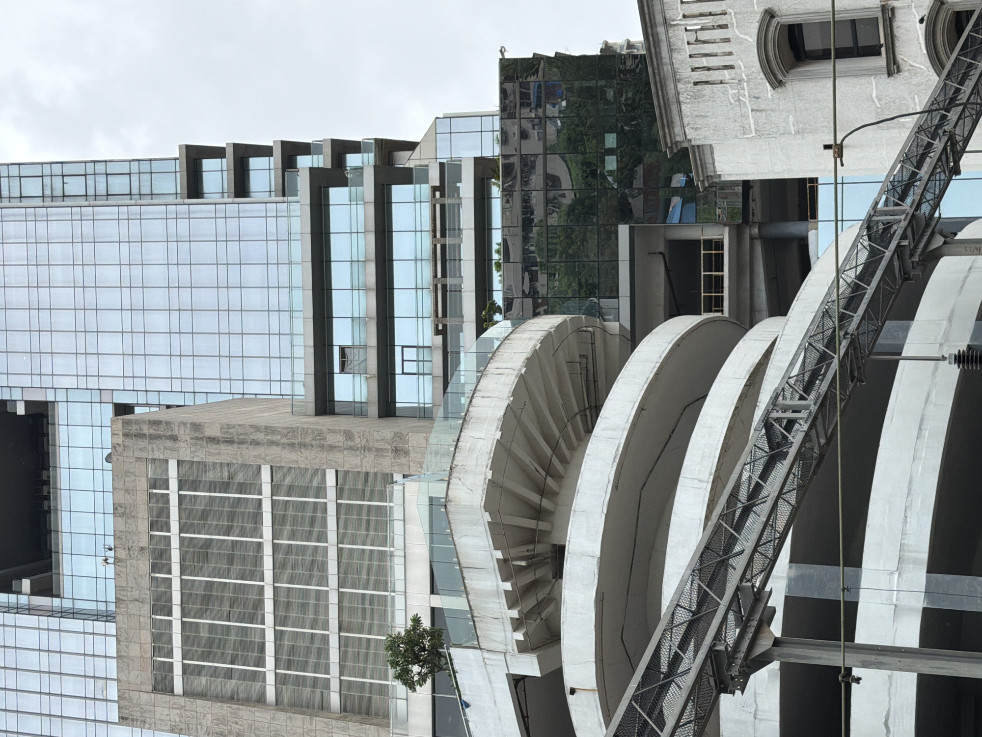 The image shows a multi-level parking structure with a prominent spiral ramp, in front of a modern glass-facade skyscraper. The parking structure is made of concrete and appears to be somewhat aged, with visible signs of wear and tear on the concrete surfaces. A metal walkway or bridge spans across part of the parking structure. The skyscraper in the background has a grid-like pattern of blue-tinted glass panels. The sky is overcast, suggesting a cloudy day. The scene is likely located in a dense urban area, given the scale and proximity of the buildings. The image does not contain any people or animals. The time of day appears to be daytime. The location is in Mumbai, India.