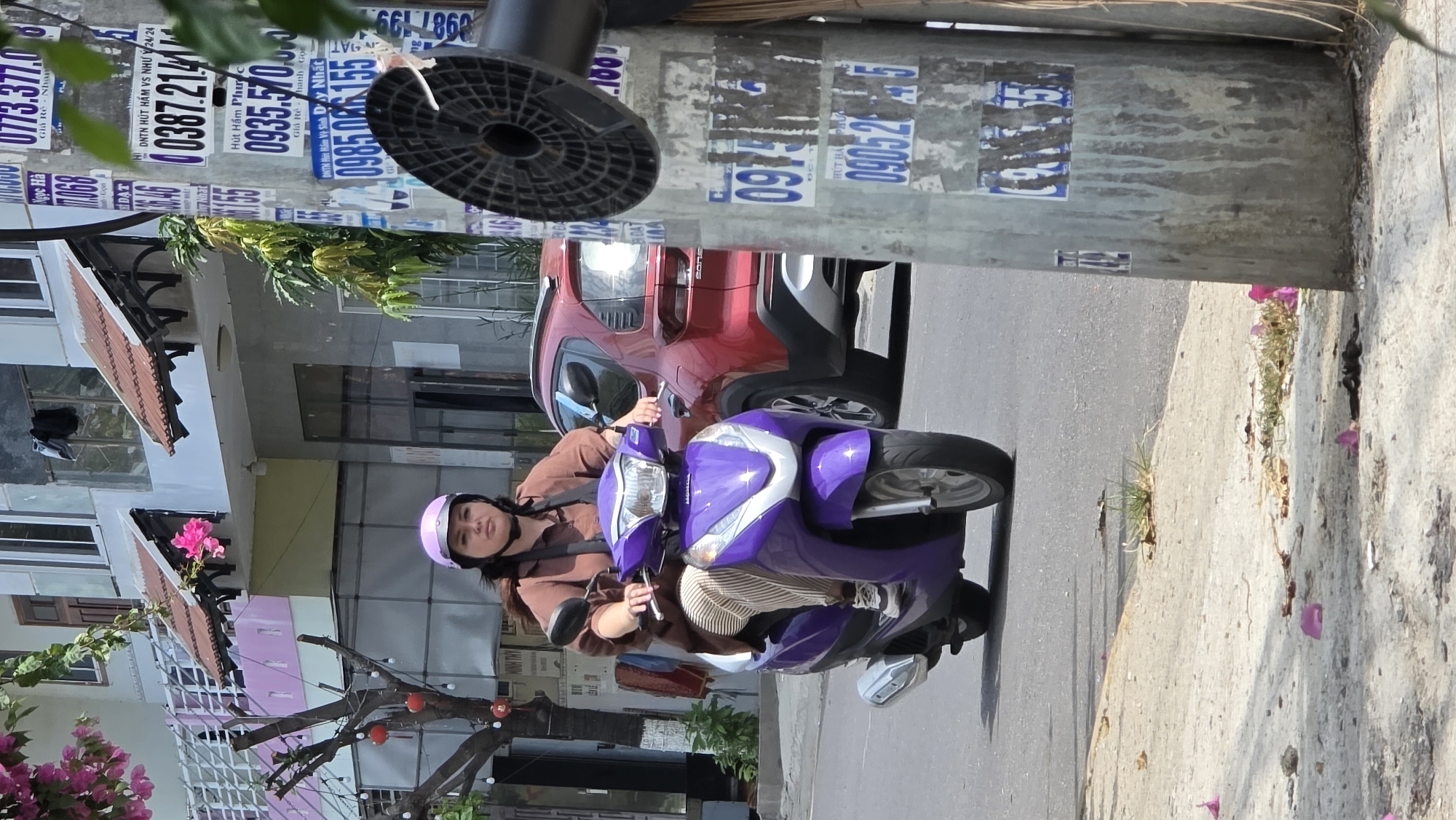 A young woman wearing a pink helmet drives a purple Honda scooter on a street in Bac Nha Trang Ward, Vietnam. She wears a brown button-down shirt. The scooter is in motion, its front wheel nearly touching the sidewalk, which is marked with scattered pink flowers. A red car is parked behind the scooter. A light pole covered with faded advertisements stands to the scooter's right. Buildings and a tree with pink flowers are visible in the background.
