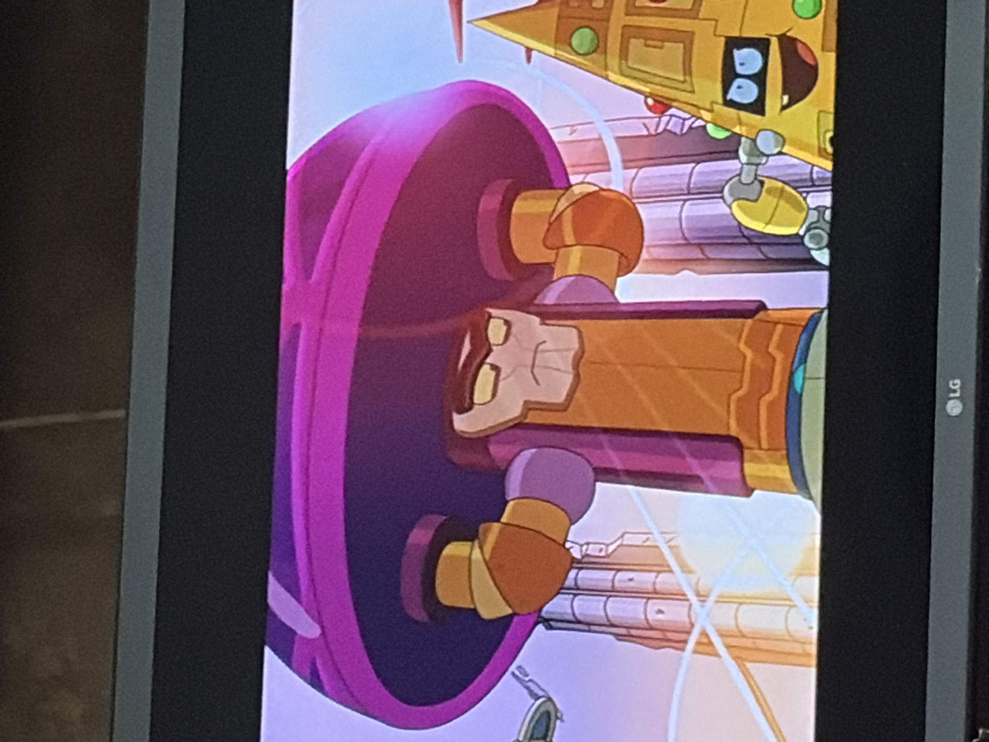 The image shows a scene from an animated show, featuring two characters and a city backdrop. On the left, a large, imposing figure with a purple, mushroom-shaped helmet dominates the frame. The figure is clad in orange and purple attire, with yellow and orange robotic arms. Its face is stern, with angled eyebrows and a grim expression. Behind this figure, tall, futuristic buildings rise into a pale sky. To the right, a yellow, triangular character with a face is visible. This character has large blue eyes, a wide grin, and is adorned with green circular decorations. A small, yellow robotic arm extends from it, reaching towards the central figure. The overall environment suggests an urban setting with a futuristic, possibly alien, aesthetic. The scene is captured from a television screen.