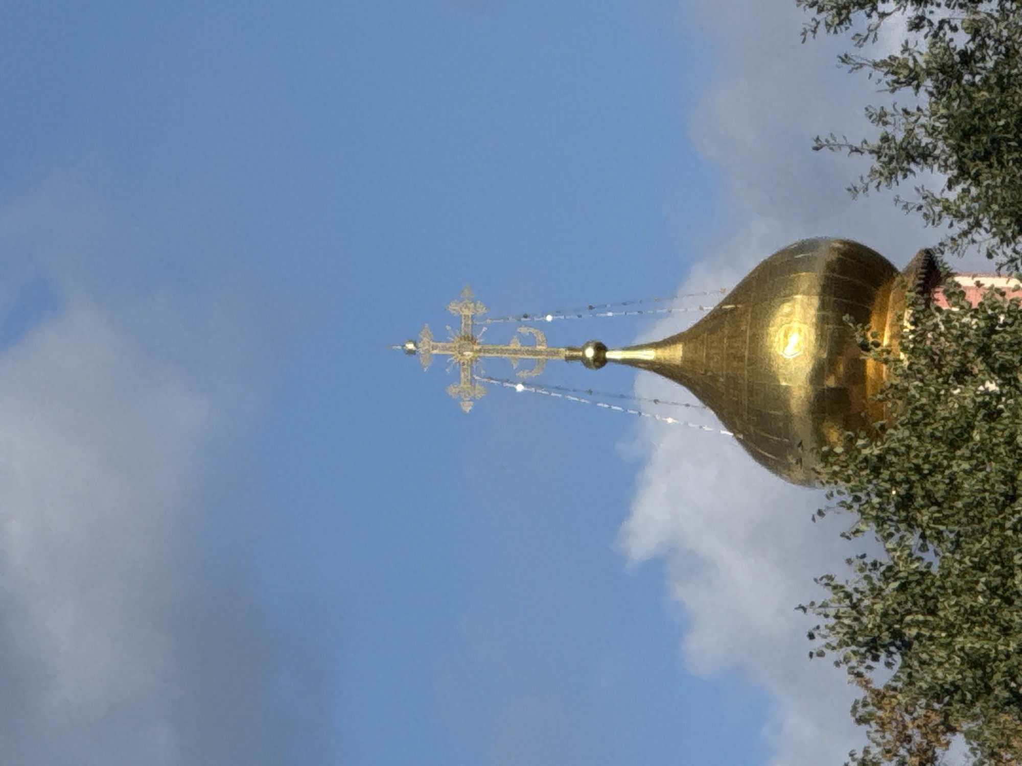 The image shows a close-up, low-angle view of the golden dome and cross of an Orthodox church, likely in Moscow, Russia, against a blue sky with scattered clouds. The dome is polished and reflects the sunlight, with a finial topped by an ornate, multi-tiered cross. Decorative chain-like elements hang from the cross. The top of a red-and-white striped structure is visible below the dome, and the upper branches of a dense green tree are in the foreground, partially obscuring the lower portion of the church building. The lighting suggests it is daytime, possibly late morning or afternoon, given the shadows and the brightness of the dome. There are no people or animals visible in the frame.