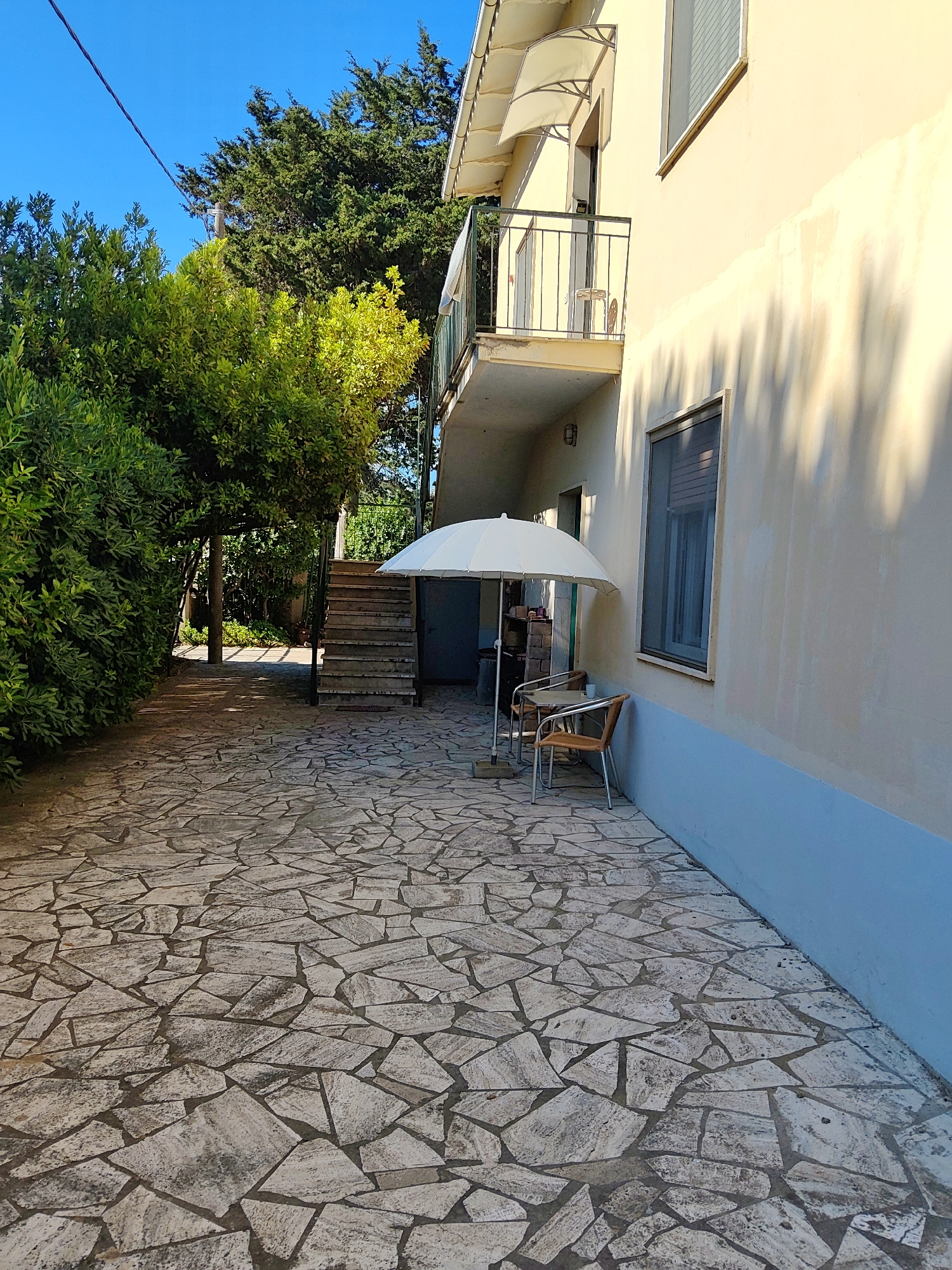 A paved walkway of irregular flagstones leads past lush green foliage and a building with a pale yellow facade. Two chairs and a small table are arranged under a large white umbrella. Stairs with metal railings ascend to a balcony on the building's exterior. The scene appears to be a residential courtyard in Campiglia Marittima, Italy, bathed in the warm sunlight of a clear day, casting dappled shadows on the ground and wall.