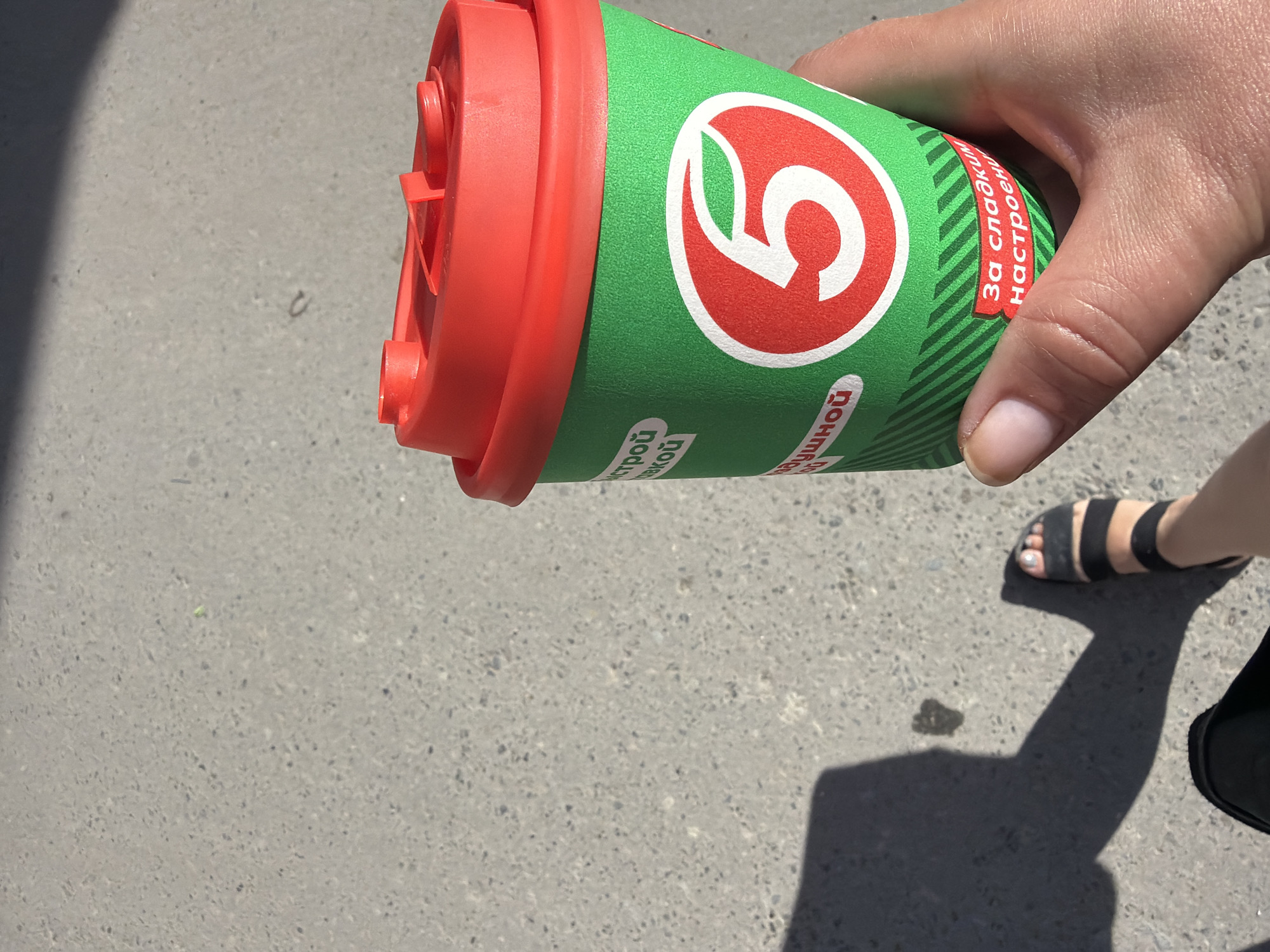 A person's hand holds a green paper coffee cup with a red lid. The cup has a white logo with a red border and a number "5" inside. There is text in Russian on the cup. The cup is held in front of a grey asphalt surface. A shadow of a leg and foot wearing a black sandal is cast on the ground. The scene is outdoors, likely on a sidewalk or street.