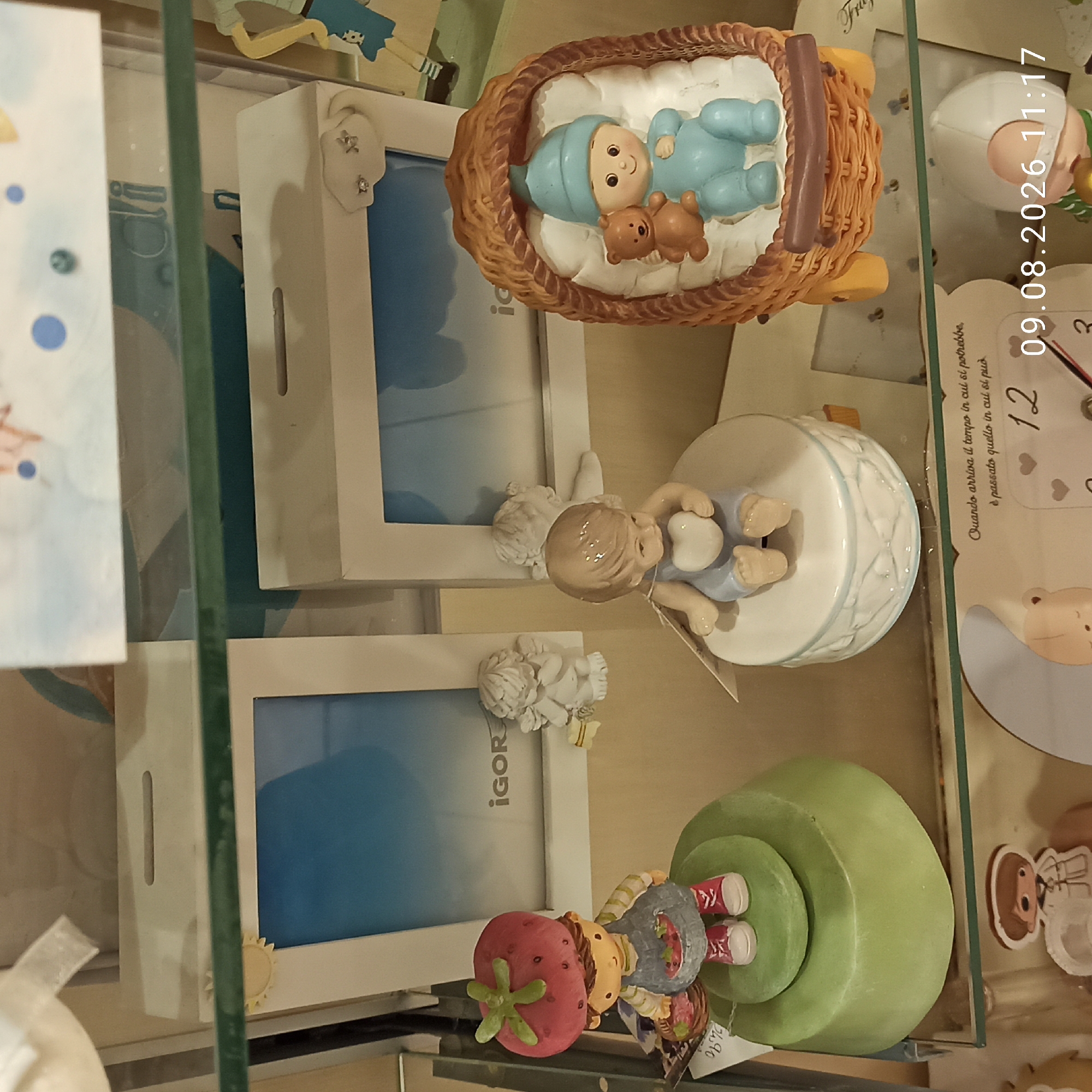 The image shows a display of baby-themed decorative items on shelves. In the foreground, on a green tiered stand, is a figurine of a strawberry girl holding a basket. Next to it is a round ceramic box with a baby figurine holding a heart. To the right, a woven crib holds a baby figurine with a teddy bear. Behind these, on the shelves, are two white photo frames with blue inserts, one featuring the name "IGOR". Small angelic figurines are placed near the photo frames. At the bottom of the image, a clock face is visible with the date "09.08.2026 11:17" superimposed. The setting appears to be a retail store, possibly a gift shop or a baby boutique. While the specific location within Molfetta, Italy is not provided, the context suggests a commercial establishment in that city.