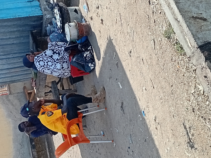 The image depicts an outdoor scene in Sharada, Nigeria. Three individuals are present. In the foreground, a male wearing a yellow collared shirt, dark trousers, a dark baseball cap, and tan tactical boots sits on an orange plastic chair, his right hand resting on his chin, looking toward the right. Behind him, a second male in a dark blue shirt is seated on a wooden bench, holding a dark object, possibly a phone, to his left ear. To the right, a female wearing a white and black patterned dress and a dark headscarf sits on a red plastic crate. She also holds a dark object to her left ear.
Adjacent to the female, a food preparation area is set up on the ground. This includes a metal charcoal stove with a round griddle on top, containing multiple light-colored items, possibly food. Several metal pots and containers, one stacked on another, are positioned near the stove. A black plastic bag is visible on the ground beside the red crate. The ground itself is dirt and sand, covered with scattered debris including wood chips and small pieces of paper. In the background, there is a building with a light-colored wall on the left and a corrugated metal wall, partly painted blue, on the right. A concrete curb or barrier runs along the lower right edge of the frame. Sunlight indicates daytime, casting distinct shadows across the ground.