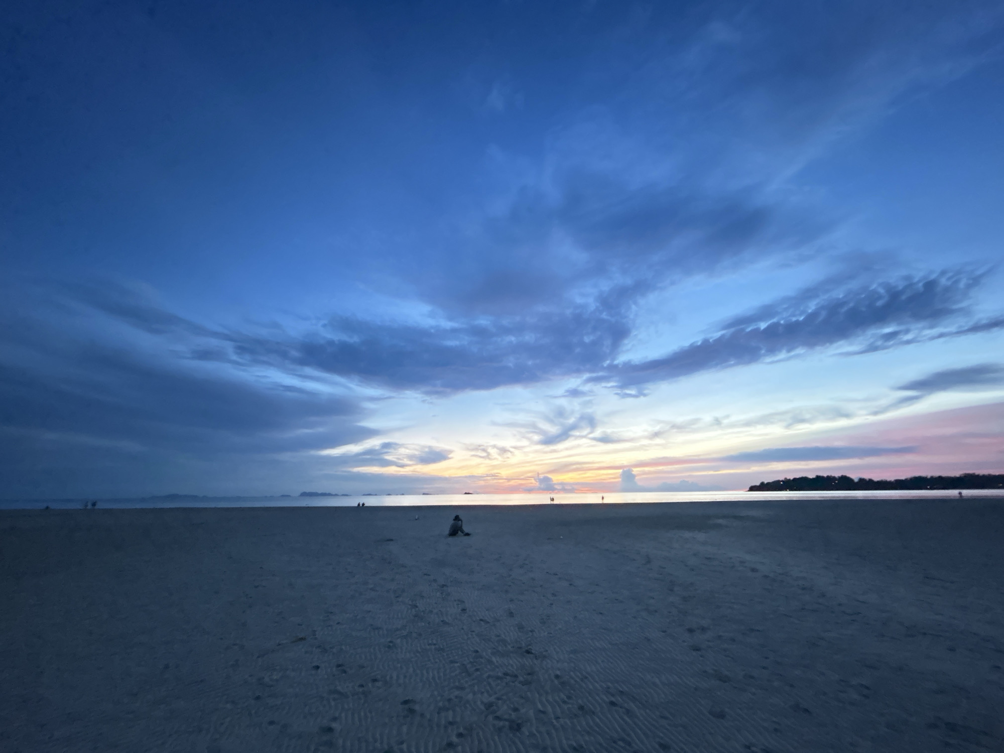 An aerial wide shot captures a beach scene at dusk on Koh Phangan, Thailand. The sky transitions from a deep blue at the top to a lighter blue and pink/orange hue near the horizon. Wispy clouds are scattered across the sky. The foreground is a sandy beach with visible ripple patterns from the receding tide. A solitary person is seated in the middle of the beach, facing the water. In the distance, the sea is calm and meets the horizon. A faint line of trees or landmass is visible on the far right of the horizon. Several other figures are visible as silhouettes along the distant shoreline, some appearing to be walking. The overall mood is tranquil, illuminated by the soft light of sunset.
