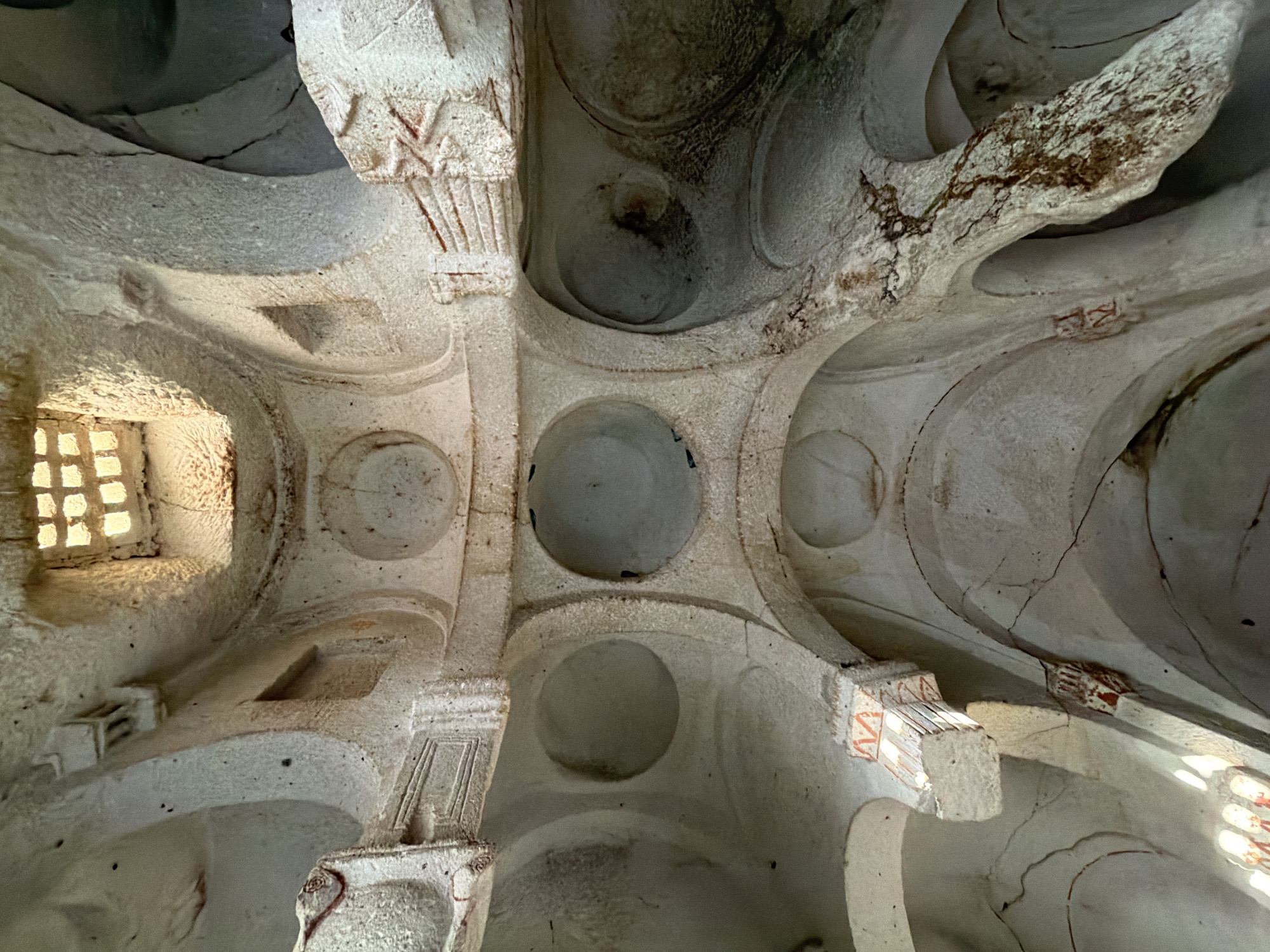This media file captures an upward-angled interior view of a rock-cut structure, strongly characteristic of a cave church or monastery found in Ürgüp, Cappadocia, Turkey.
The primary subjects are the architectural elements carved directly from the soft, light-colored stone or tuff. Four robust pillars, adorned with stylized geometric patterns and remnants of reddish pigment, anchor the central space. These pillars support a ceiling featuring multiple circular, dome-like indentations, creating a textured and segmented overhead surface. Arched connections extend between the pillars and the surrounding walls.
Natural daylight illuminates the interior, streaming in through at least two windows. A prominent rectangular window on the left, with a distinct grid-like pattern, casts bright light onto the rugged wall surfaces. Another window on the right side also allows light into the ancient space.
No people or specific activities are depicted, emphasizing the timeless and static nature of the architecture. The scene conveys a sense of historical depth and unique craftsmanship. The time of day appears to be daytime, as indicated by the natural light from the windows.
Notable details include the weathered condition of the stone, showing cracks, textured erosion, and dark stains, possibly from moisture or smoke accumulation over centuries. The reddish pigment on the carved pillar details hints at former decorative schemes. On a small, block-like protrusion near the ceiling, a faint red mark resembling a stylized "K" or similar letter/symbol is visible. Other visible markings are abstract geometric patterns painted in reddish hues on the column capitals.