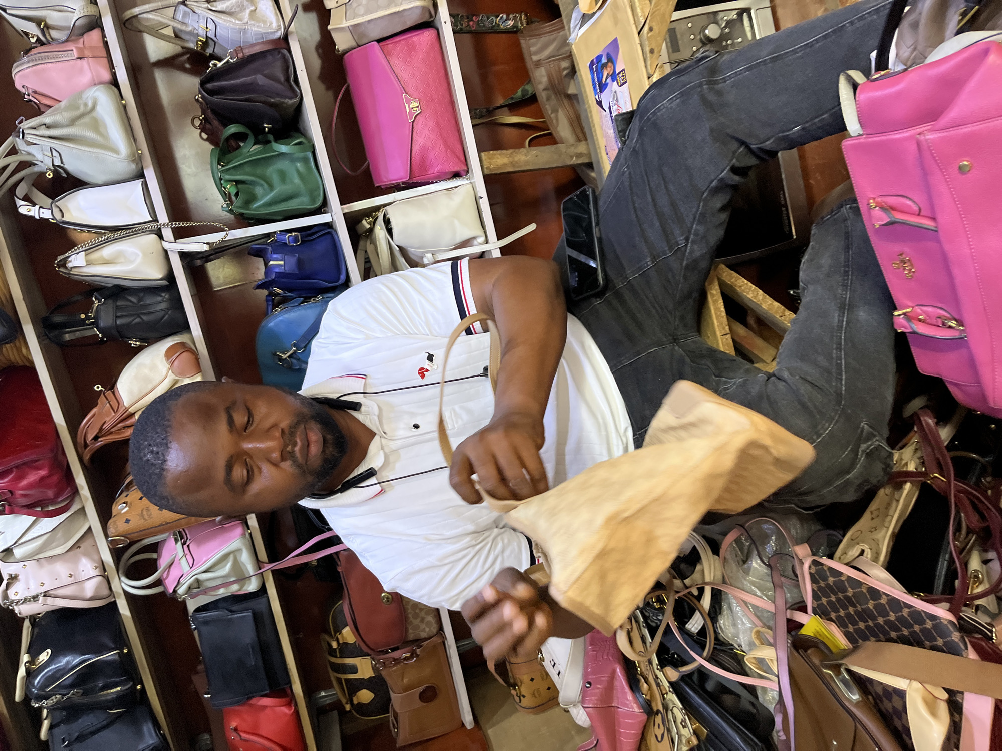 A man with dark skin and short black hair is sitting in front of a wall of shelves stacked with handbags. He is wearing a white polo shirt with red and blue trim on the collar and sleeves, and dark wash jeans. He has an earpiece in his left ear. The man is holding a tan-colored fabric bag in his hands, and appears to be inspecting it. A black smartphone is resting on his lap. The shelves behind him are filled with a variety of handbags in different colors, sizes, and styles, including black, pink, red, white, green, and brown. Some of the bags appear to be designer brands. The environment appears to be a retail space, likely a handbag store or market stall, possibly located in Abuja, Nigeria. The lighting is somewhat dim, suggesting an indoor setting.