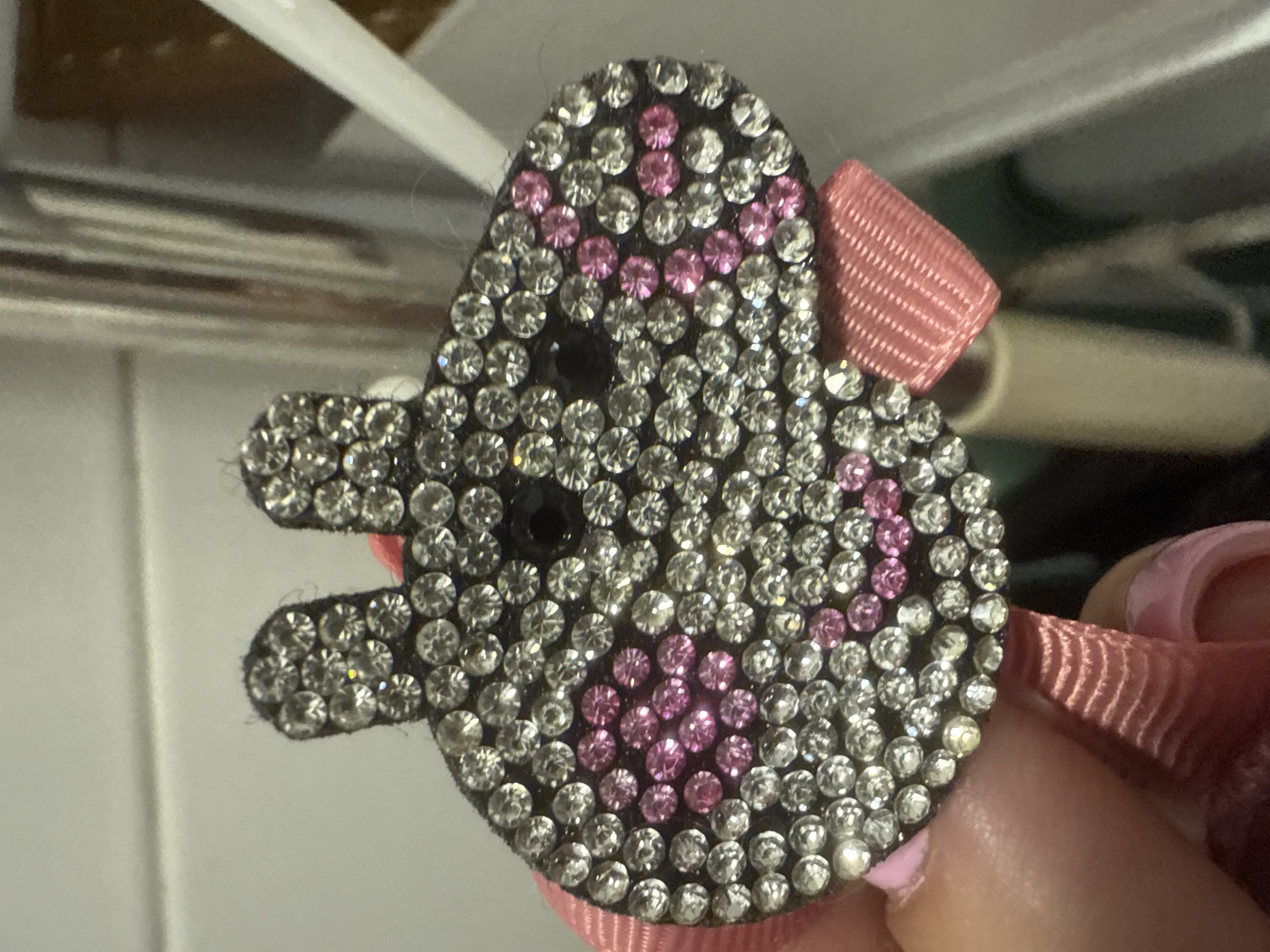 The image shows a close-up of a handmade hair clip shaped like a pig's face, adorned with clear and pink rhinestones. The pig has two round ears at the top, two black eyes, and a snout formed by pink rhinestones. A pink ribbon is attached to the bottom of the pig's face, creating a bow. The clip is being held by a person's hand, with a fingernail painted pink visible in the lower left corner. The background is blurred, showing a light-colored, tiled wall and what appears to be a metal or plastic object to the right. The image does not provide any information about the location in Kroyanskoye, Russia.