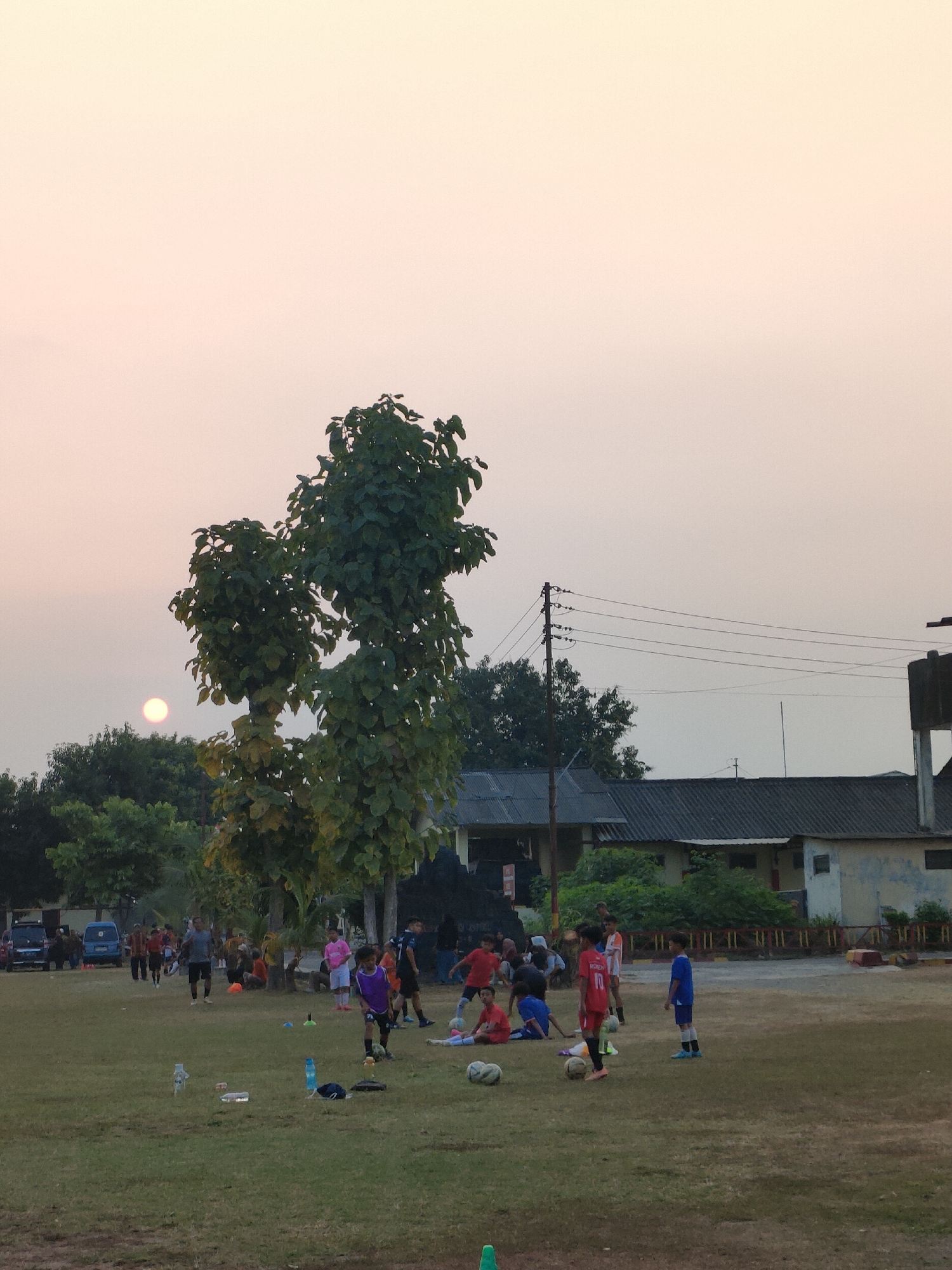 This is a photograph of a soccer practice taking place on a grassy field in Banguntapan, Indonesia. The image captures a group of children and adults, some of whom are wearing soccer uniforms, engaged in activities related to the sport. Several soccer balls are visible on the field, along with training cones. In the background, there are buildings, trees, and a utility pole with overhead wires. The sky is a soft, pale orange, suggesting either sunrise or sunset, with a faint sun visible in the upper left portion of the frame. The overall atmosphere appears to be casual and recreational.