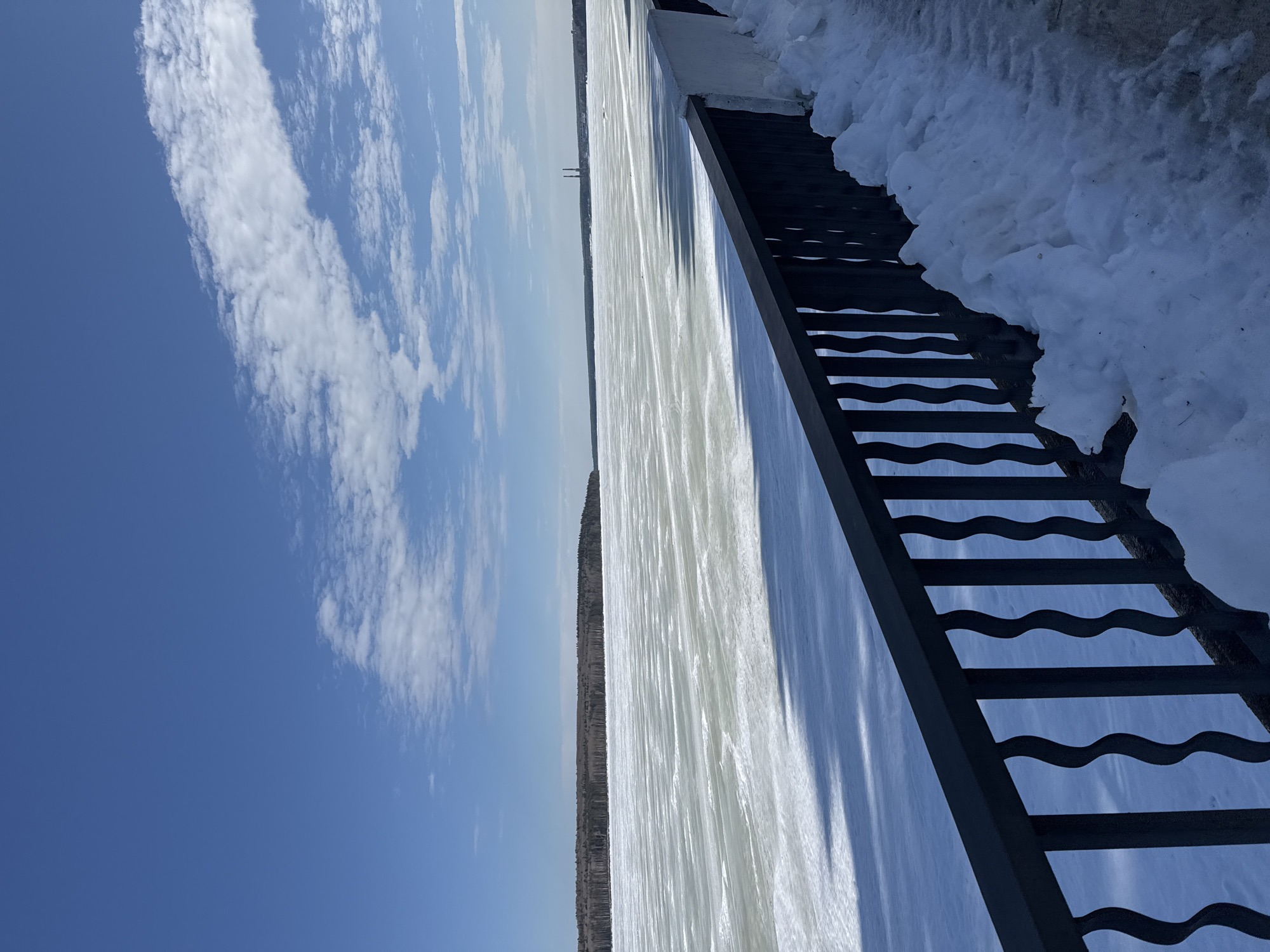 The image depicts a wide, frozen expanse identified as the Votkinsk Reservoir in Chaikovsky, Russia, observed under a clear blue sky with scattered white cumulus clouds. In the immediate foreground, a dark-colored metal balustrade featuring undulating vertical pickets runs horizontally across the lower frame. To the right of the balustrade, a white concrete parapet is partially obscured by a substantial accumulation of dirty snow and ice on what appears to be a paved pedestrian area. Long, distinct shadows from unseen foreground structures are cast across the textured, snow-covered surface of the frozen reservoir. The distant shoreline is demarcated by a dense, dark-barked forest. On the far right horizon, two prominent industrial chimneys or stacks are visible. No human or animal figures, or overt actions, are present in the scene.