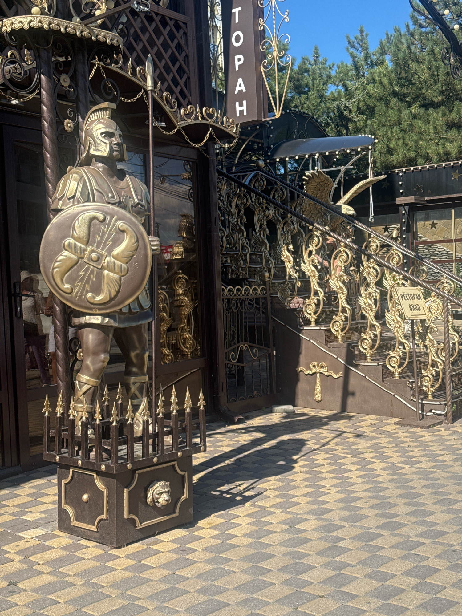 The image shows an ornate entrance to a building. In the foreground, a life-sized bronze statue of a Roman soldier stands guard. The soldier is depicted wearing a helmet, armor, and sandals, holding a spear in his right hand and a shield in his left. The shield has a decorative emblem featuring stylized lightning bolts. The statue stands on a decorative pedestal with a lion's head motif.
Behind the statue, a dark brown metal gate and a staircase with a golden railing are visible. The railing is elaborately decorated with intricate scrollwork and what appear to be winged figures. A sign with Cyrillic text, "РЕСТОРАН ВХОД" (Restaurant Entrance), is attached to the railing. The building's facade features dark brown doors and windows. The ground is paved with a checkered pattern of grey and yellow rectangular tiles. In the background, green trees and a clear blue sky are visible.
The overall aesthetic suggests a themed establishment, possibly a restaurant or entertainment venue, with a focus on classical or historical motifs. The sunlight casts strong shadows on the tiled ground, indicating a bright, sunny day. The location is identified as Anapa, Russia, which is a resort city on the Black Sea coast.