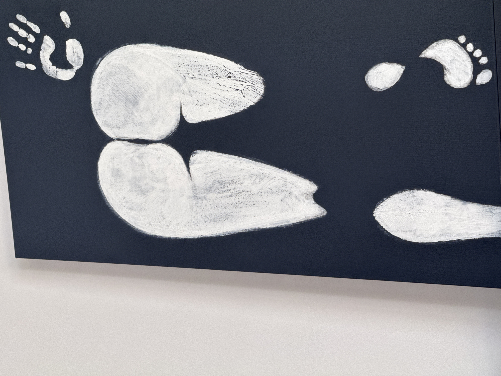 A painting on a dark canvas depicts white handprints and footprints. At the top, a handprint is visible, with individual fingers and a thumb clearly defined. Below this, a large, abstract shape formed by two connected footprint-like impressions suggests buttocks. Further down, two more footprints are depicted, one above and to the left of the other, oriented as if walking. The painting is displayed indoors, leaning against a plain white wall. The image does not contain any information about the city of Budva, Montenegro.