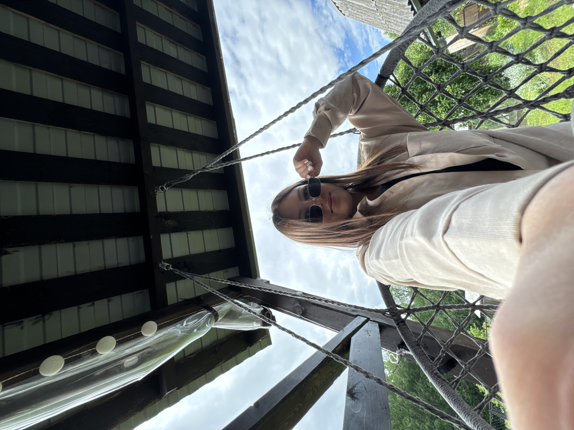 A young woman is seated in a rope hammock suspended from a wooden structure. She is wearing sunglasses and a light-colored jacket, and is looking at the camera. The hammock appears to be outdoors, possibly on a porch or in a backyard. The background shows trees and a cloudy sky. The overall impression is one of a casual, relaxed moment. The location is not specified in the image or crops.
