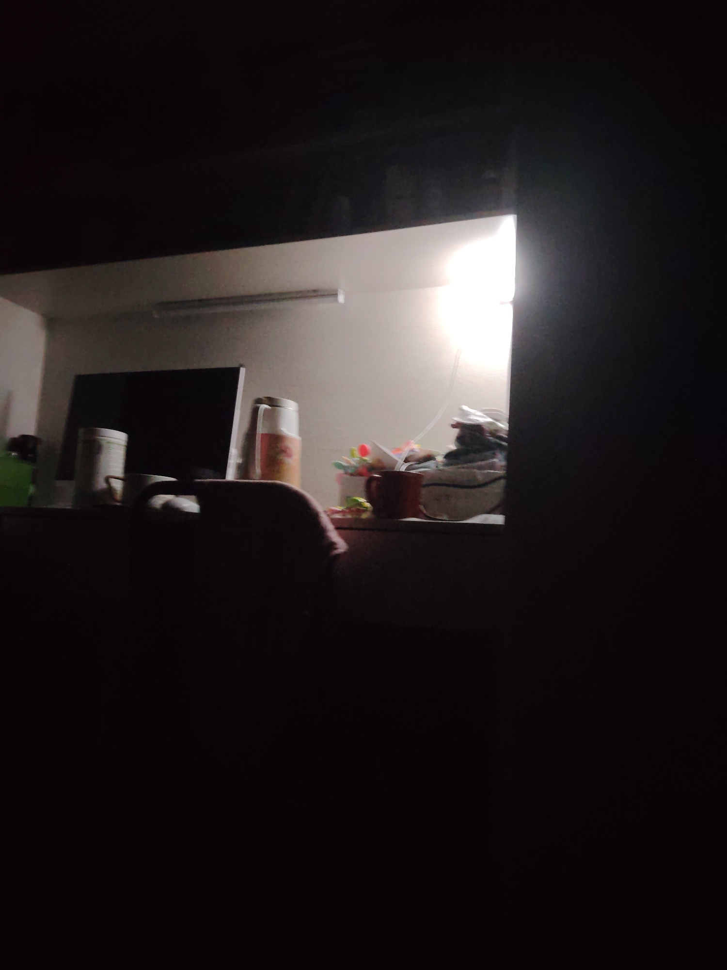This is a dark, low-light, slightly blurry photograph of a desk in what appears to be a room in Termez, Uzbekistan. The desk is cluttered with various objects. To the left of the frame, there is a dark computer monitor. In front of the monitor are two cylindrical containers, one white and one a lighter shade with a pattern. A white mug is also visible. To the right of the monitor, a thermos with a patterned design is standing upright. Further to the right, there is a collection of items including a red mug and some indistinct colorful objects that might be pens or flowers. The background appears to be a wall with a light fixture or window emitting a bright light from the upper right portion of the scene. The overall impression is of a workspace at night, illuminated by a desk lamp or overhead light. There are no people visible in the photograph.