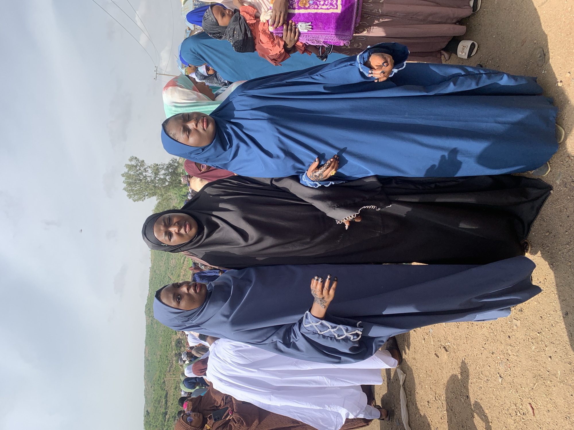 The image shows a group of women in Naraguta Hausawa, Nigeria. Three women are in the foreground, wearing different colored hijabs: blue, black, and blue again from bottom to top. These women are in line next to each other. The hands of the blue hijabs show henna and beaded embellishments. Other women can be seen in the background, wearing different colored hijabs and clothing. The ground is dirt.