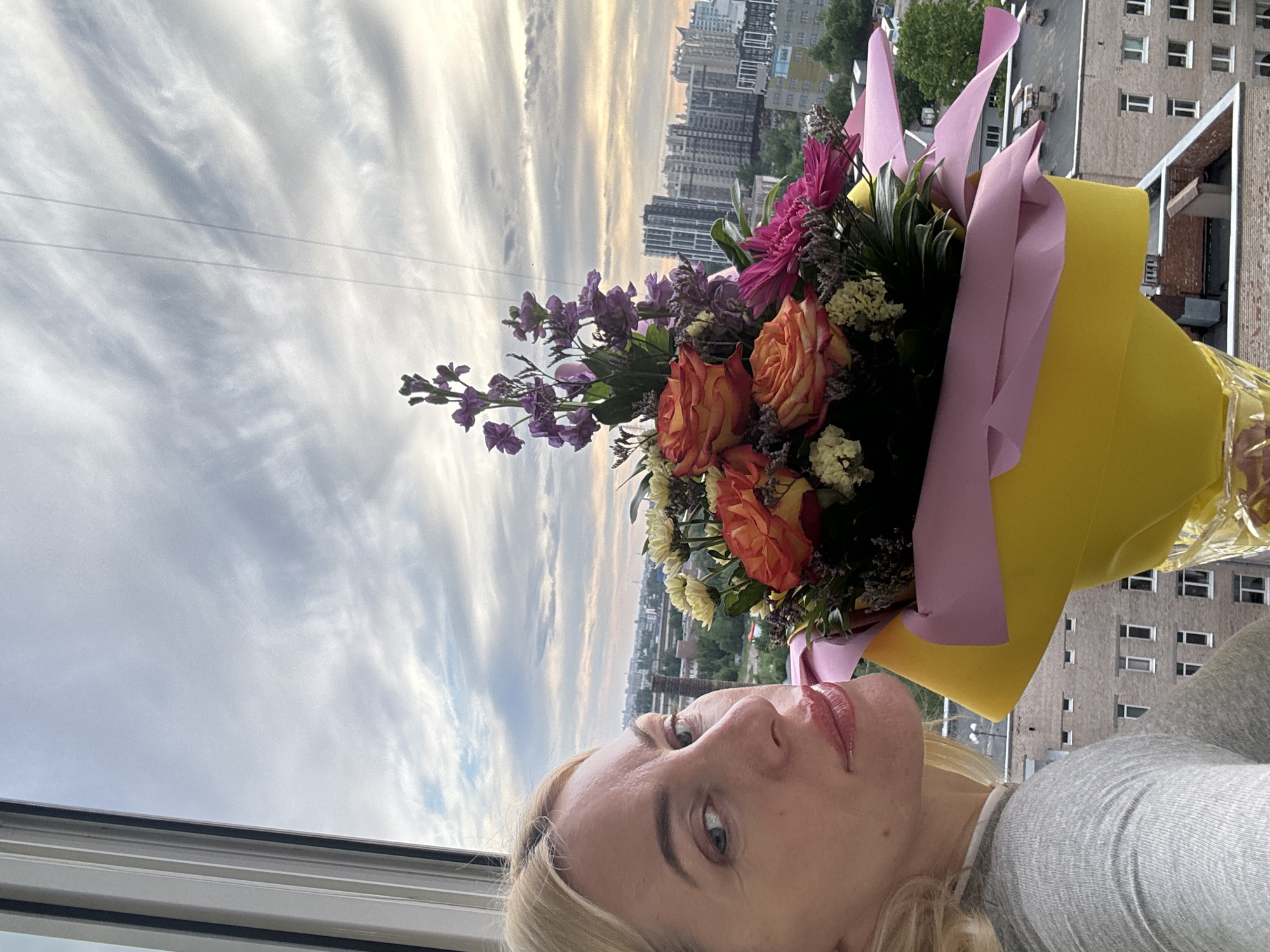 A self-portrait image captures a blonde woman holding a vibrant bouquet of flowers in front of a city skyline, viewed from a high vantage point during what appears to be late afternoon or early evening.
In the foreground, the woman's face, from her forehead to her chin, is visible on the left side of the frame. She has light-colored eyes, and her lips are adorned with light-colored lipstick. Her expression is neutral, and she looks directly at the camera. She wears a light grey top.
To her right, a large, colorful bouquet of flowers is prominently displayed. It features bright orange and red roses, tall stalks of purple stock flowers, pink chrysanthemums or gerberas, and smaller white and yellow filler flowers, interspersed with green foliage. The bouquet is wrapped in bright yellow paper with soft pink paper or ribbons providing accent.
The background reveals an expansive urban landscape seen through a window. Numerous tall apartment or commercial buildings stretch across the horizon. Beyond the city, the sky is filled with sweeping, thin cirrus clouds, bathed in the warm, soft light of the sun nearing the horizon, creating a gentle glow. Faint power lines are visible against the sky in the upper portion of the image.
The overall scene conveys a serene moment, featuring a personal detail against a dramatic urban sky. No discernible text is visible in the image.