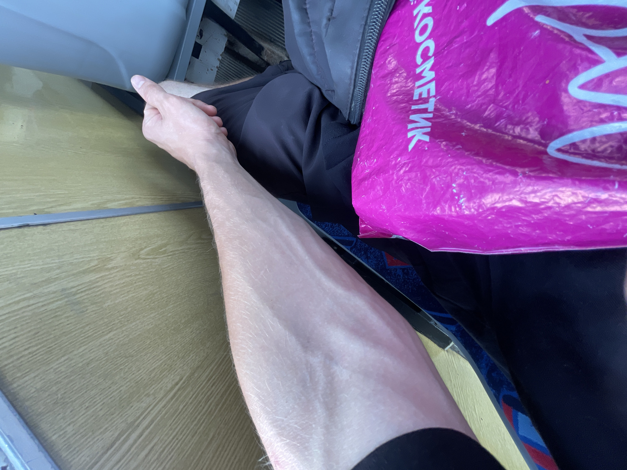 The image shows a close-up view of a person's forearm and hand, which is resting on their lap. The person is wearing black shorts and a black jacket is visible. In the foreground, a bright pink plastic bag with white lettering is present. The background appears to be the interior of a vehicle, with a wood-paneled wall and a grey vertical element. The scene suggests the person is seated, possibly in a bus or train, given the visible interior elements. The location of Novokuznetsk, Russia, is not discernible from the visual information provided in the image.