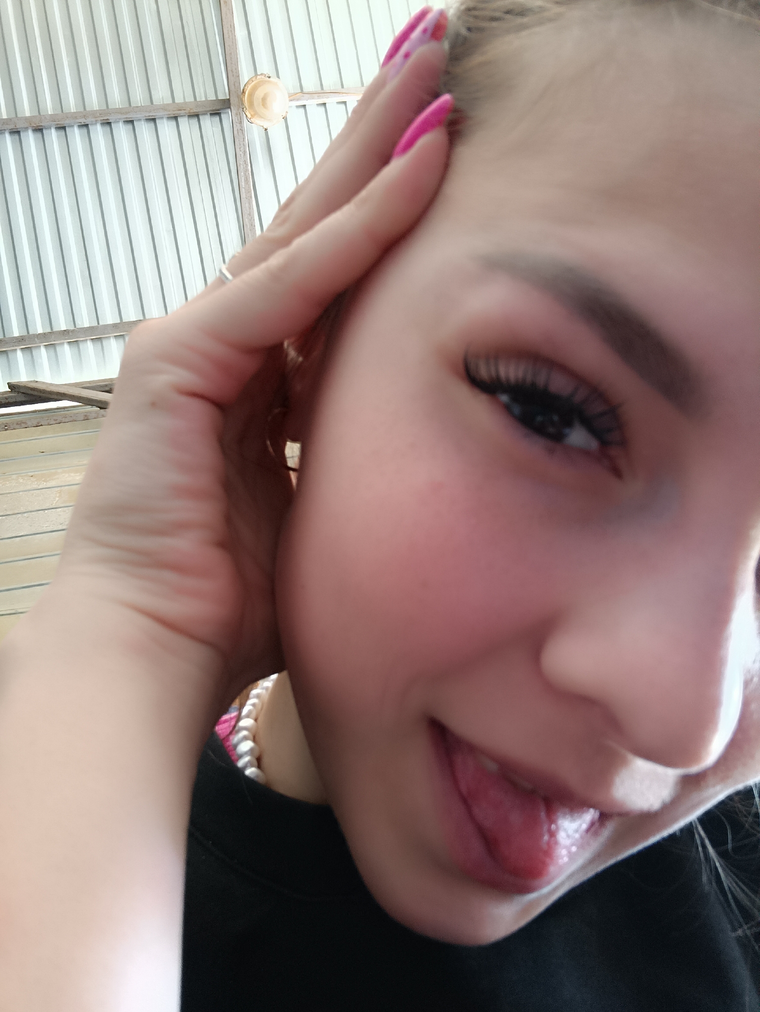 A close-up, slightly angled image shows a person with fair skin and long, dark eyelashes, sticking their tongue out. The person has a pearl necklace and is wearing a black shirt. Their hand is near their head, revealing pink nails with designs. The background includes a corrugated metal ceiling and a light fixture. The location is Unknown, Unknown.