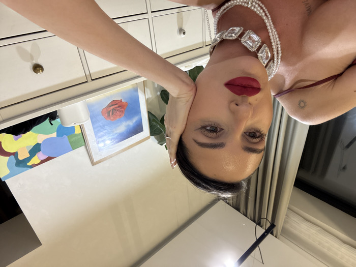 An indoor scene in a room located in Khimki, Russia, features a female individual positioned in the foreground, likely taking a selfie. She is looking directly towards the camera with a serious expression, her left hand resting against her left cheek. Her dark hair is pulled back, and she has visible makeup including winged eyeliner, dark eyebrows, and red lipstick. She wears a dark, possibly maroon, top with spaghetti straps. Adorning her neck are a multi-strand pearl necklace and a choker featuring large, square, clear stones set in silver-toned metal. A thin silver chain is also visible. Her right arm displays a small sun-like tattoo on the upper arm/shoulder and a faint "NAIVE" tattoo lower down. Her fingernails have white tips.
The background consists of light-colored walls. To the left, a window with beige or grey curtains is visible, reflecting a linear ceiling light fixture. To the right, a white multi-drawer dresser or cabinet is present. On its surface, there is a framed print depicting a red rose floating against a cloudy blue sky, an abstract painting with various colorful shapes, a small white rectangular object, and a green-leafed plant partially visible.