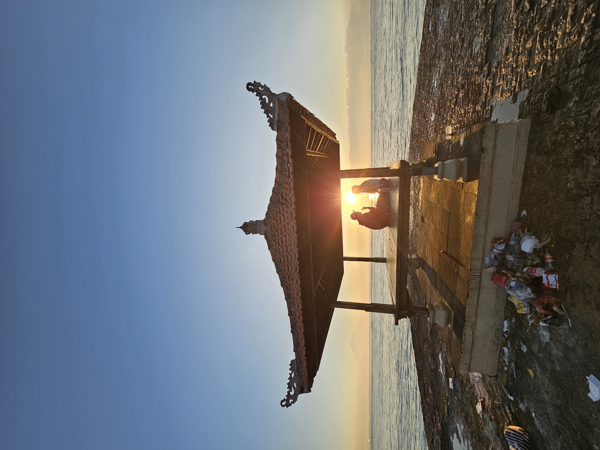 Two people are silhouetted inside a Balinese pavilion at sunrise, facing the ocean. The pavilion, with its tiled roof and ornate carvings, is situated on a rocky shore. The sun's rays are visible behind the figures, creating a bright glare. The water is calm and stretches to the horizon, where distant clouds are visible. In the foreground, the rocky ground is littered with a significant amount of trash, including plastic bottles, bags, and wrappers, suggesting a lack of upkeep. The overall scene depicts a serene natural setting marred by human-generated waste. While the exact location in Denpasar is not specified, the pavilion's architecture and the coastal environment are characteristic of many beachfront areas in Bali.