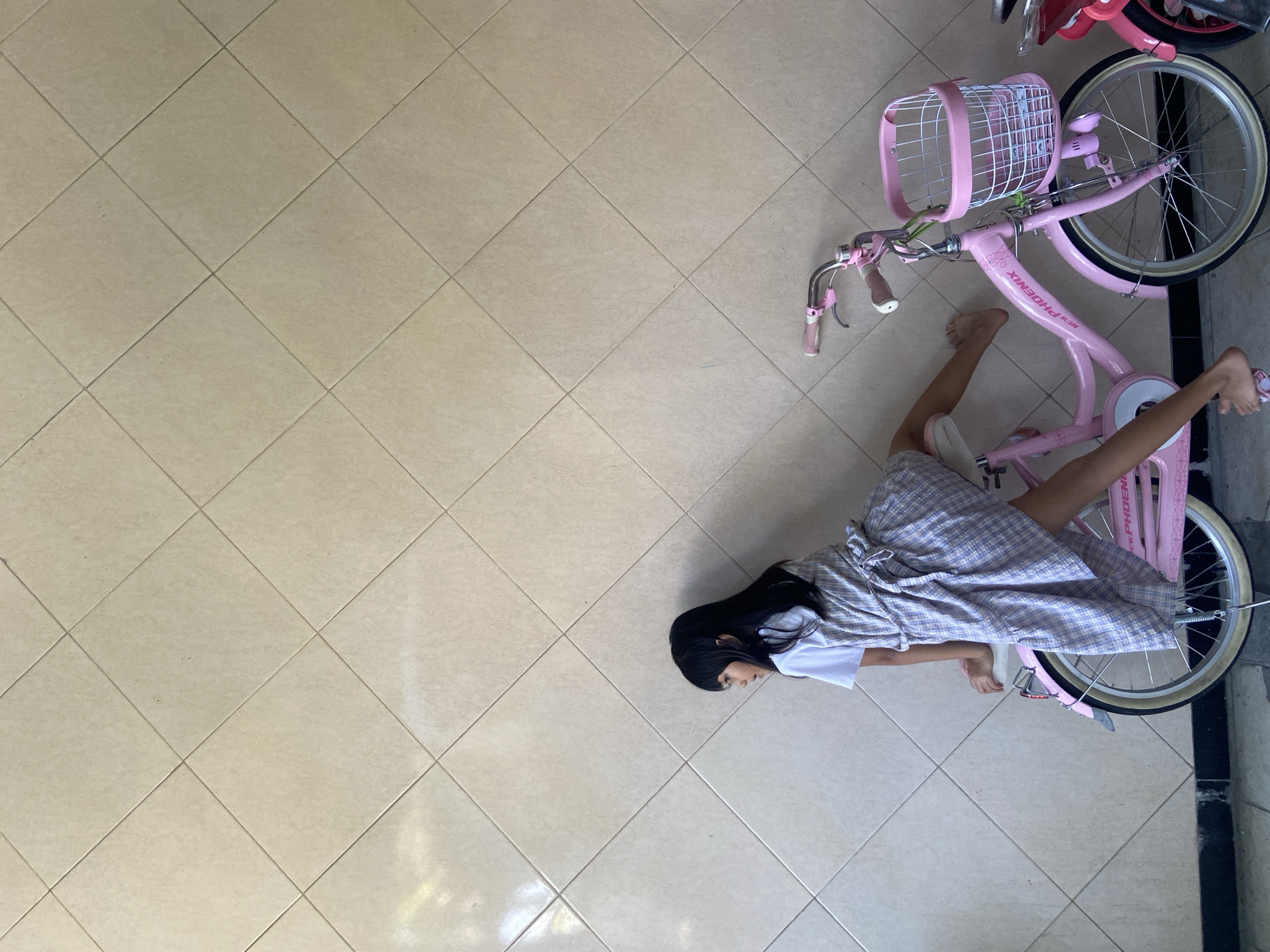 A young girl with long black hair, wearing a white t-shirt and a plaid dress, is perched on a pink bicycle. Her left leg is extended backwards, and her right leg is bent and positioned near the pedal. The bicycle has a white basket on the front. The girl is leaning against a wall covered in square, beige tiles arranged in a diamond pattern. The scene appears to be outdoors, with the ground visible at the bottom of the image showing dark and light colored pavement. A portion of another red bicycle is visible on the far right. The image has a bright, natural light quality. The location is identified as Gelam, Indonesia.